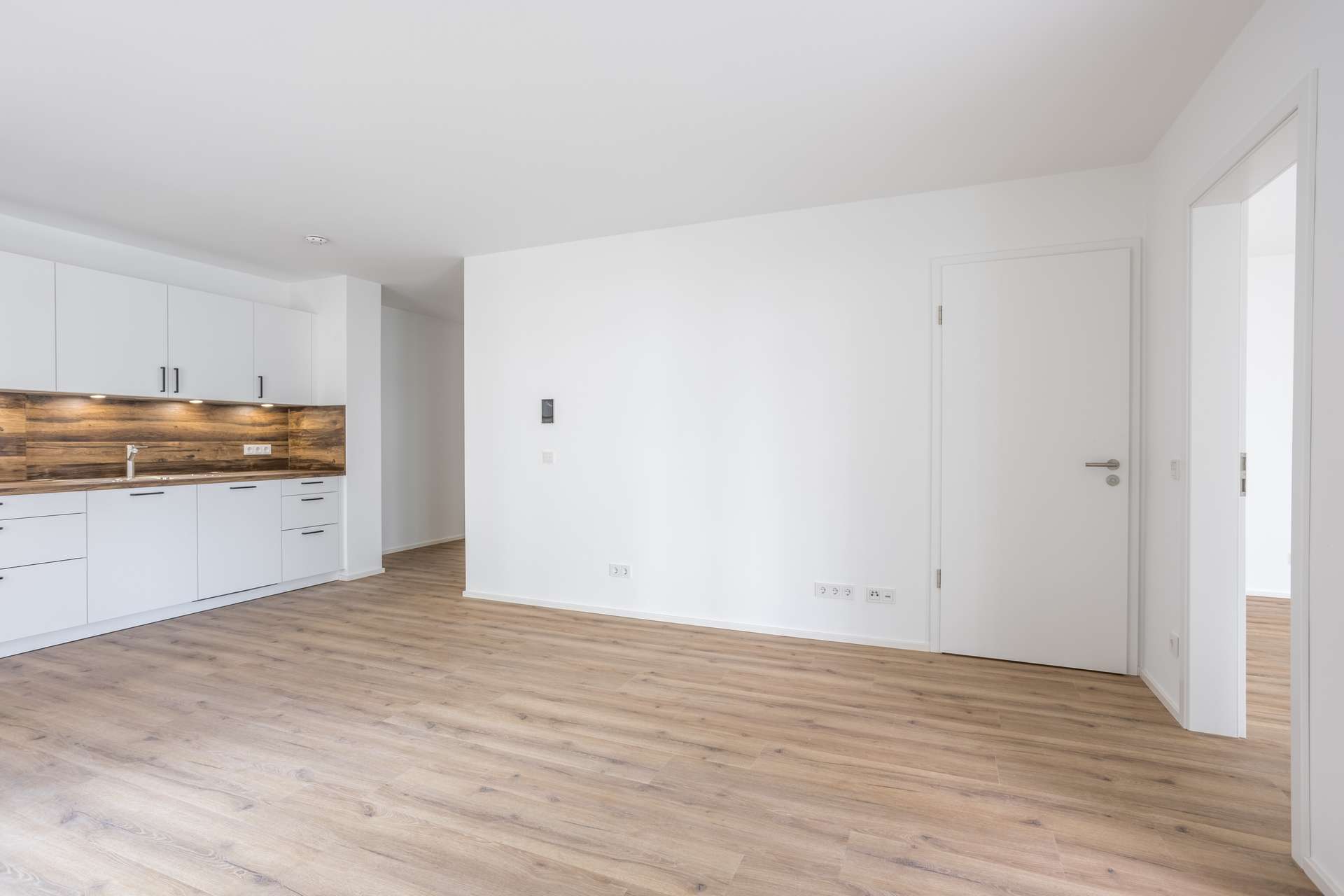 Wohn- und Essbereich Moderne, unmöblierte Wohnung mit hellem Holzboden, weißen Wänden und einer minimalistischen Küche mit weißen Schränken und einer Küchenrückwand aus Holz. Zahlreiche Türen und viel Tageslicht runden den klaren, offenen Raum ab.