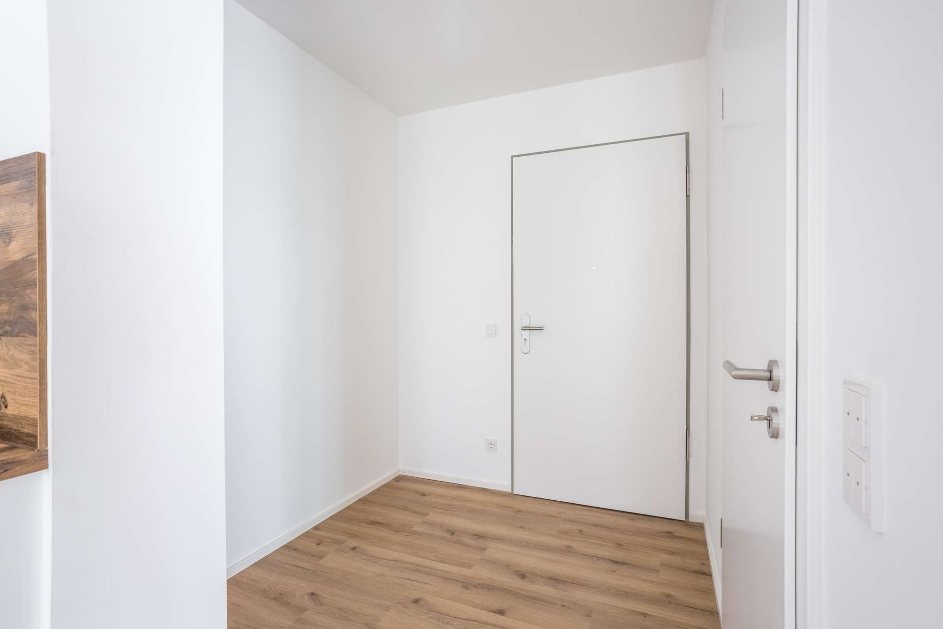 Eingangsbereich Minimalistischer Eingangsbereich mit weißen Wänden, einer geschlossenen weißen Tür mit silbernem Türgriff und hellem Holzboden. Der Raum ist leer und hell, was ihm ein klares und modernes Erscheinungsbild verleiht.