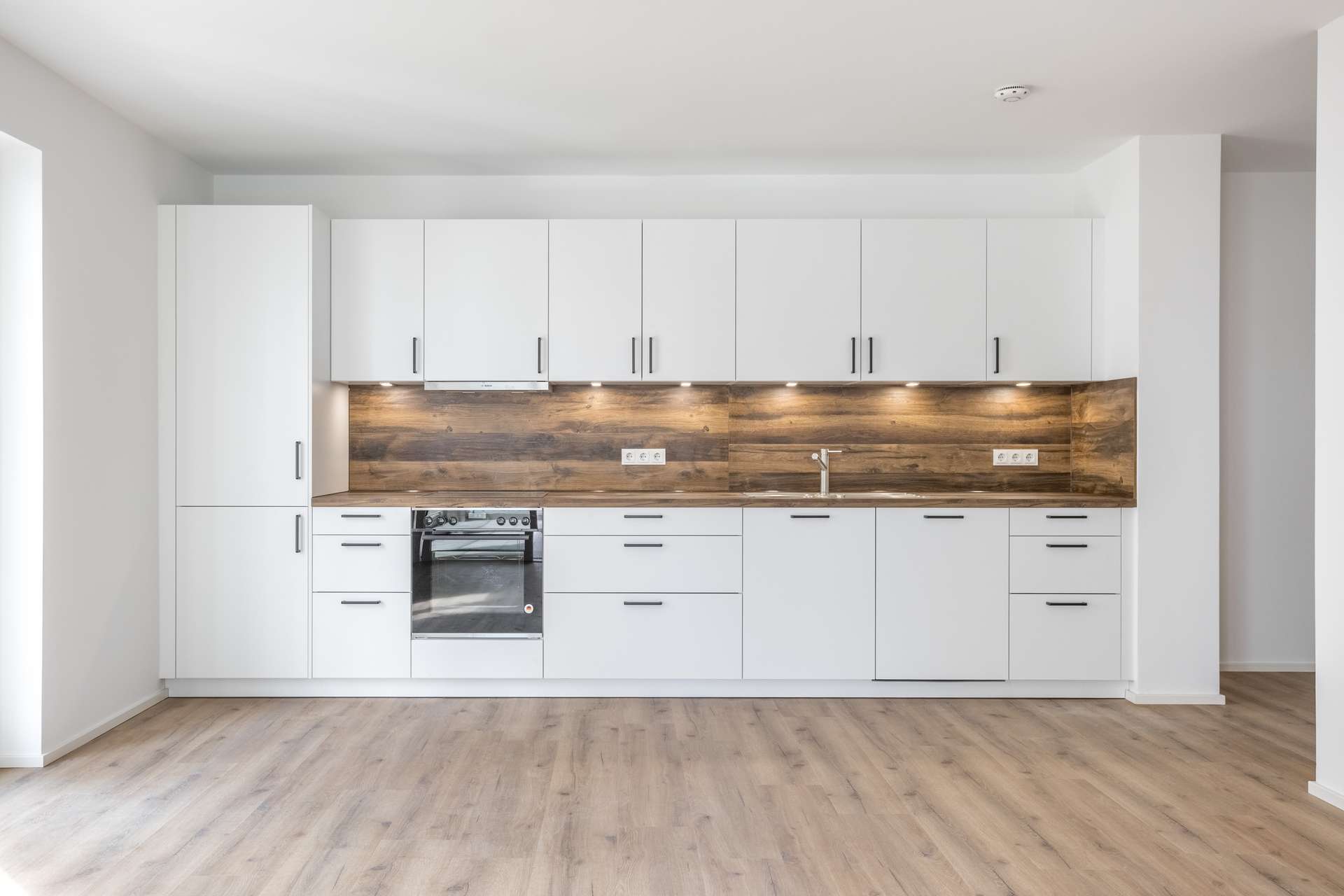 Küche Moderne Küche mit weißen Schränken, einer Küchenrückwand aus Holz, einem Einbaubackofen, einer Spüle und Unterbauleuchten. Der Boden besteht aus hellem Holz, und der Raum wirkt dank Tageslicht und Einbauleuchten hell und freundlich.