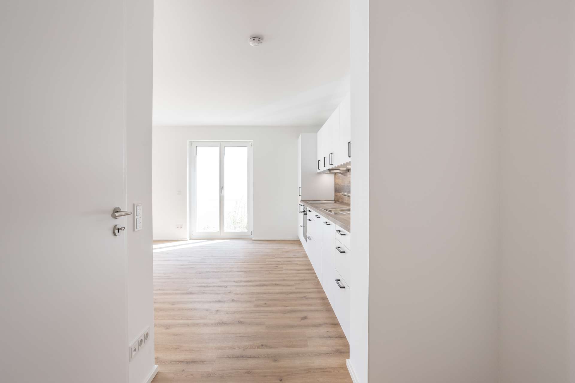 Eingangsbereich Helle, moderne Küche mit weißen Schränken, Holzboden und großen Glastüren, durch die Tageslicht hereinströmt. Das Bild wurde von einer offenen Tür aus aufgenommen und zeigt einen klaren, minimalistischen Raum.