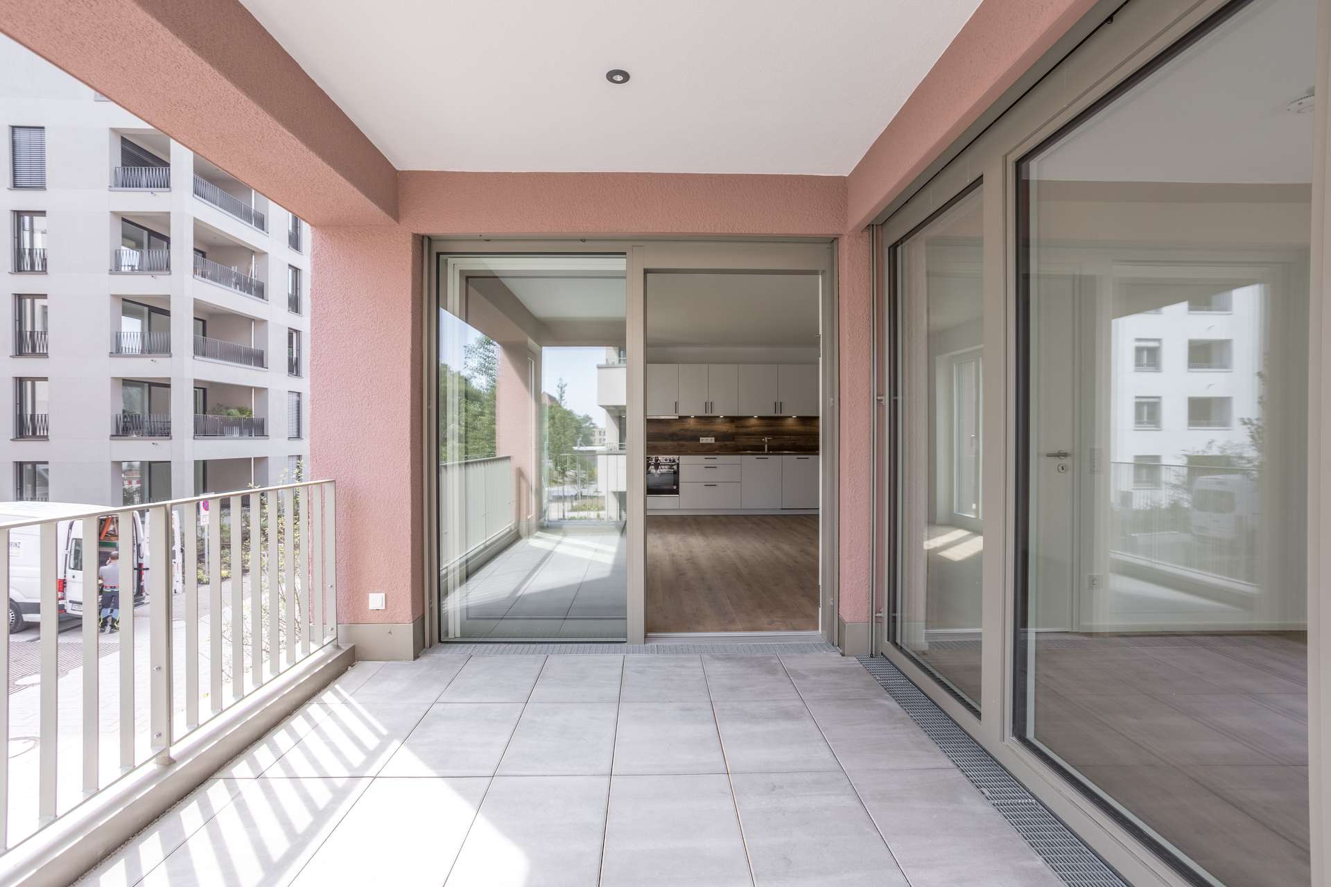 Loggia Ein moderner Wohnungsbalkon mit Fliesenboden, Glasgeländern und großen Glasschiebetüren, die zu einer offenen Küche und einem Wohnbereich mit Holzböden führen. Draußen sind Mehrfamilienhäuser zu sehen.