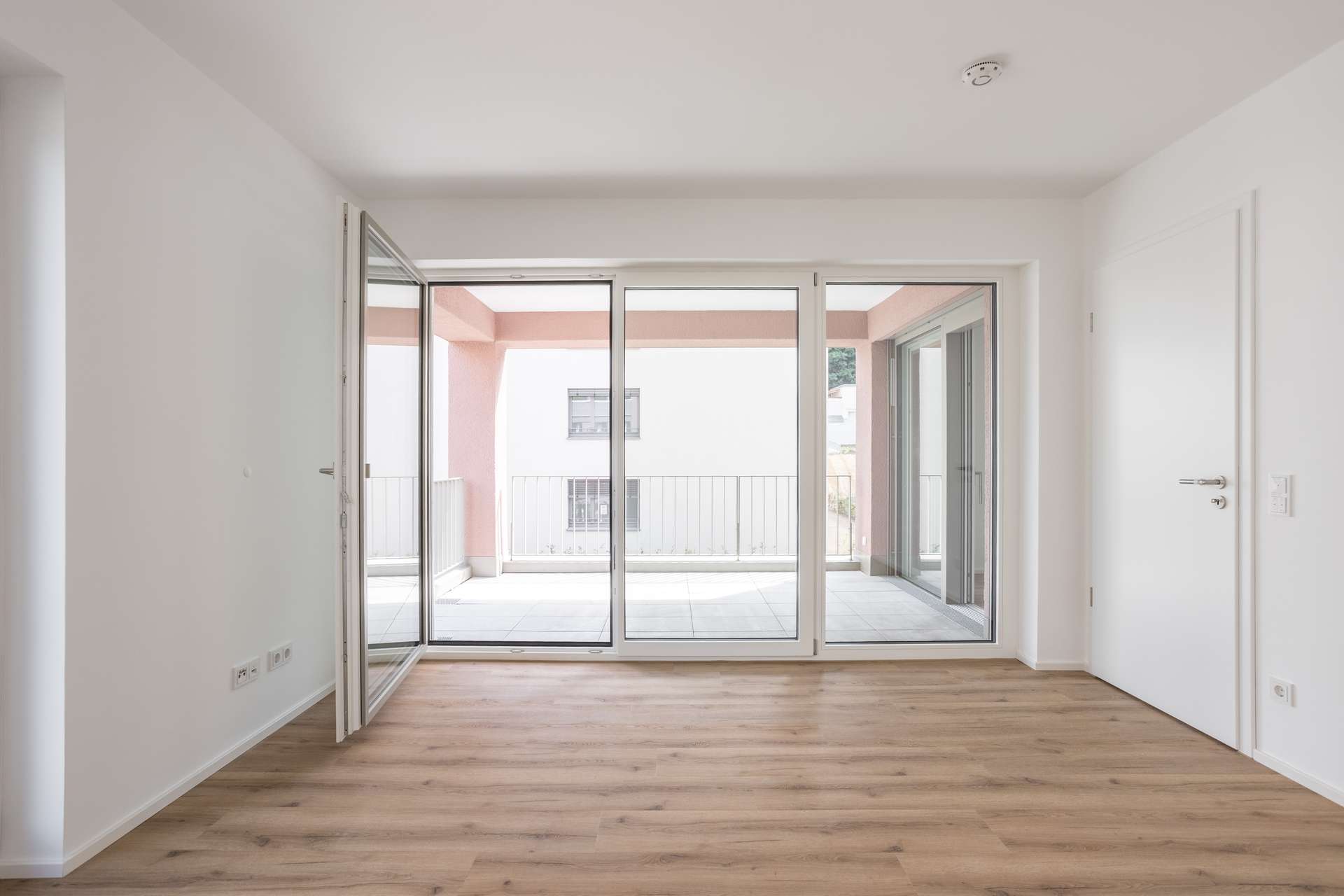 Schlafzimmer Ein heller, leerer Raum mit hellem Holzboden, weißen Wänden, großen Glastüren, die auf einen überdachten Balkon führen, und natürlichem Licht, das von draußen hereinströmt.