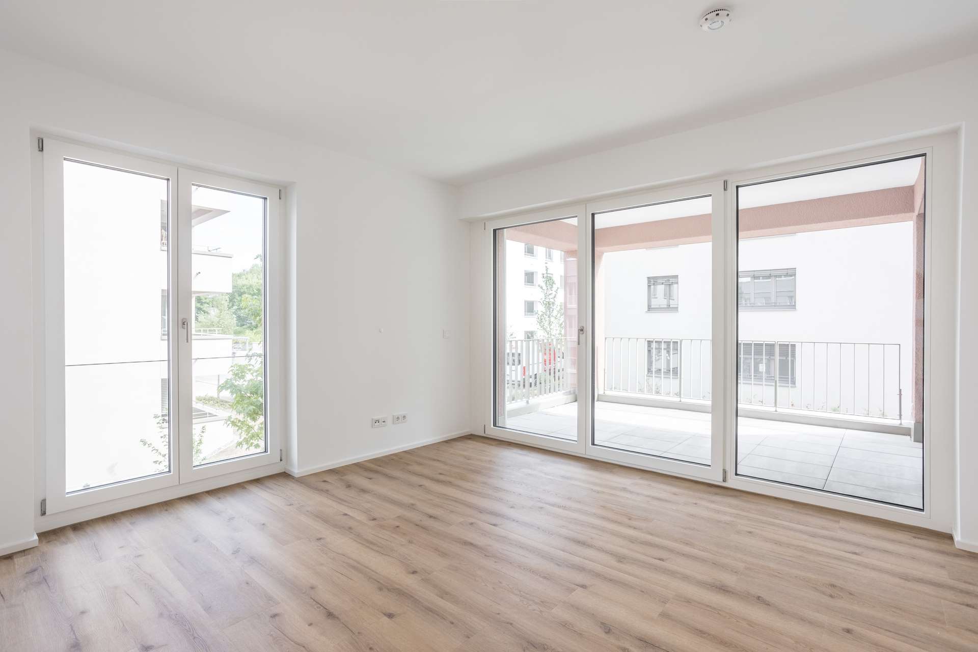 Schlafzimmer Ein heller, leerer Raum mit großen Fenstern und Glastüren, die auf einen Balkon führen. Der Raum hat weiße Wände, Holzböden und viel Tageslicht.