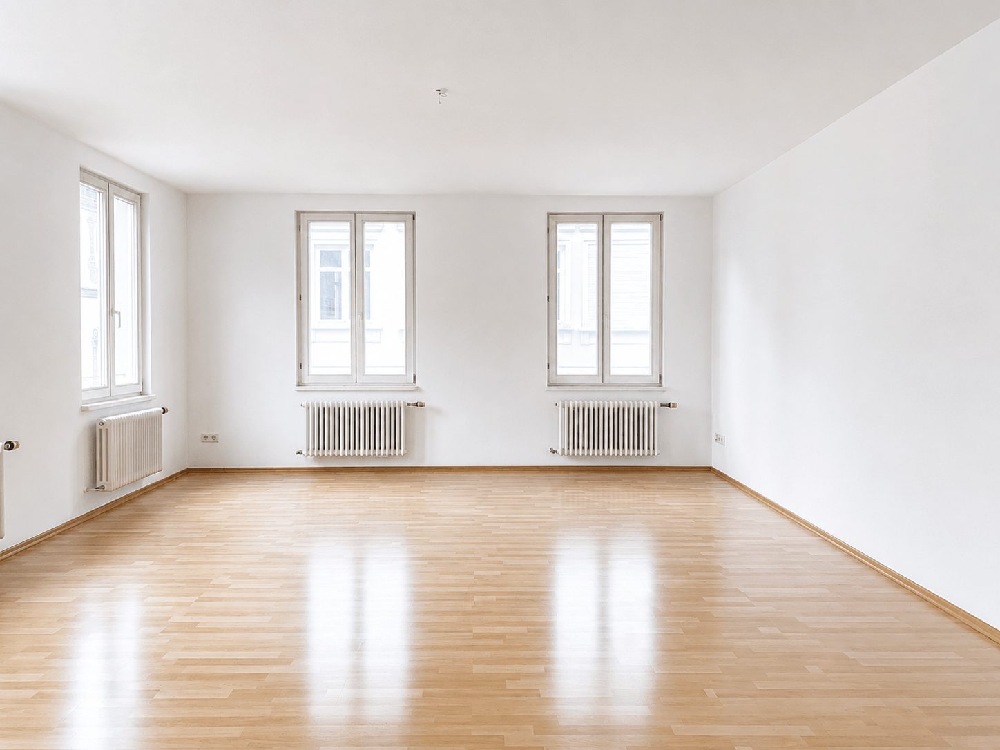 Ein leerer Raum mit hellen Holzböden, weißen Wänden, zwei großen Fenstern und Heizkörpern unter jedem Fenster. Helles Tageslicht durchflutet den minimalistischen, großzügigen Raum.