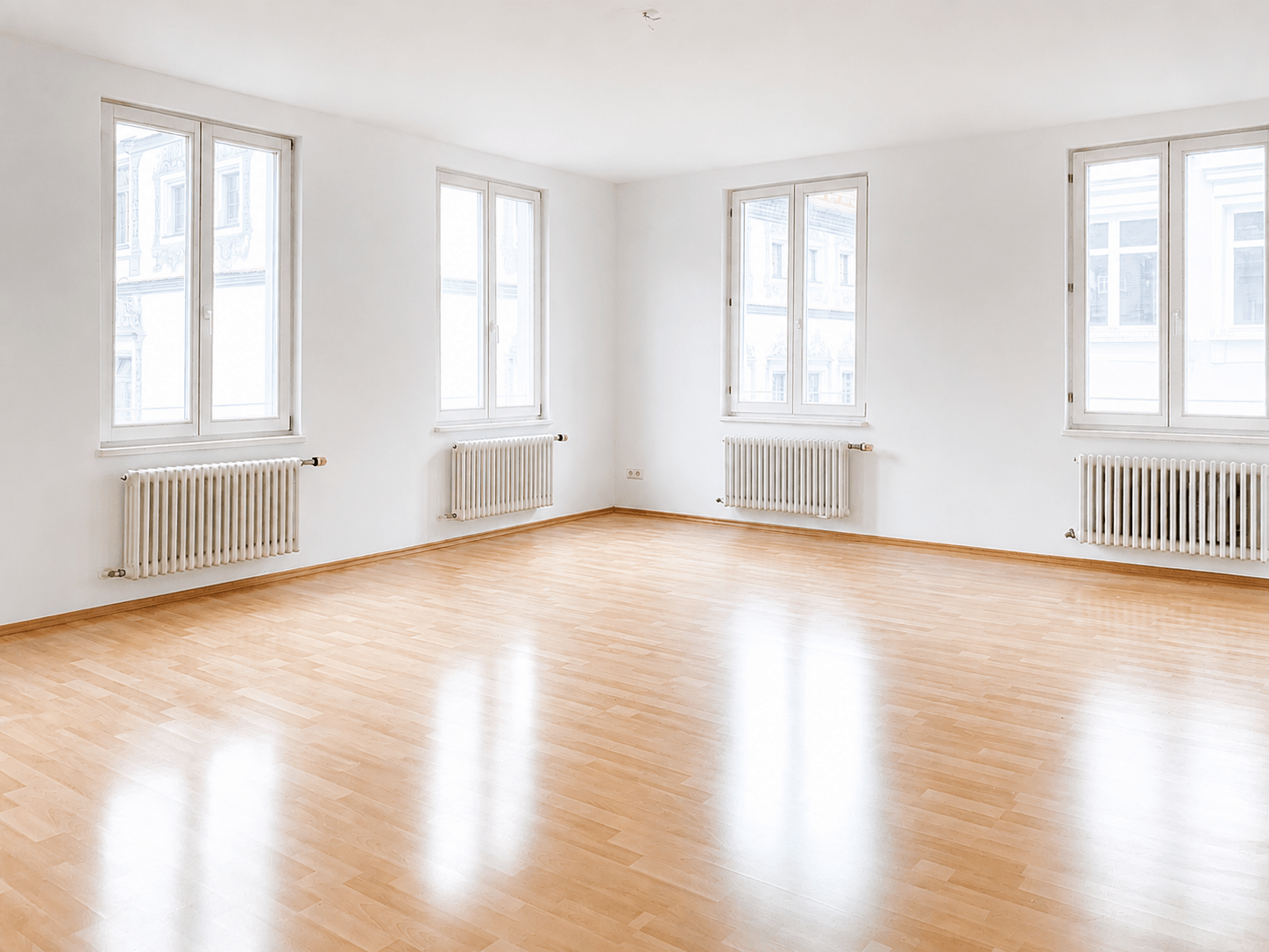 Ein heller, leerer Raum mit weißen Wänden, großen Fenstern und hellem Holzboden; unter den Fenstern befinden sich Heizkörper.