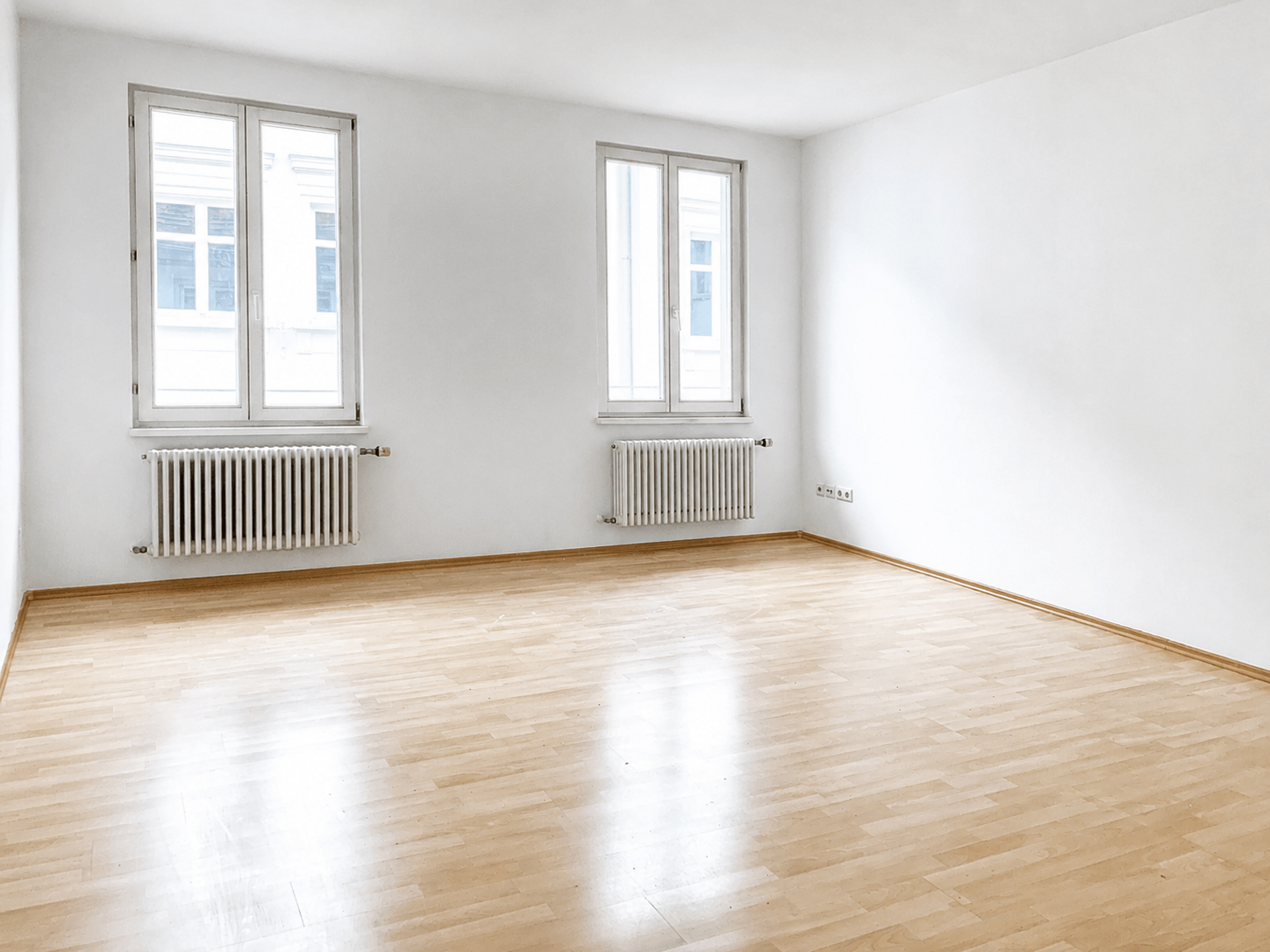 Ein heller, leerer Raum mit hellem Holzboden, weißen Wänden, zwei Fenstern und zwei weißen Heizkörpern unter den Fenstern. Der Raum ist unmöbliert, und durch die Fenster fällt Tageslicht herein.