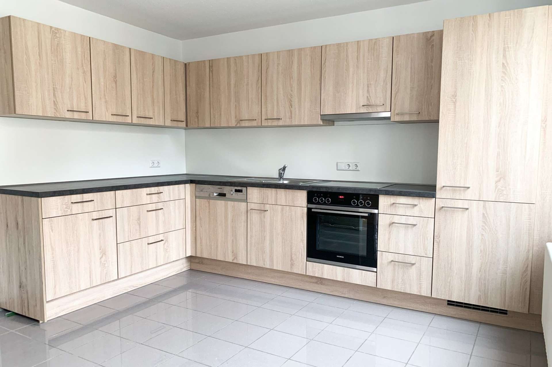 Moderne Küche mit hellen Holzschränken, schwarzer Arbeitsplatte, Einbaubackofen, Kochfeld, Geschirrspüler, Spüle und Fliesenboden. Minimalistisches Design mit reichlich Stauraum, neutralen Farben und ohne sichtbares Küchengeschirr oder Dekoration.