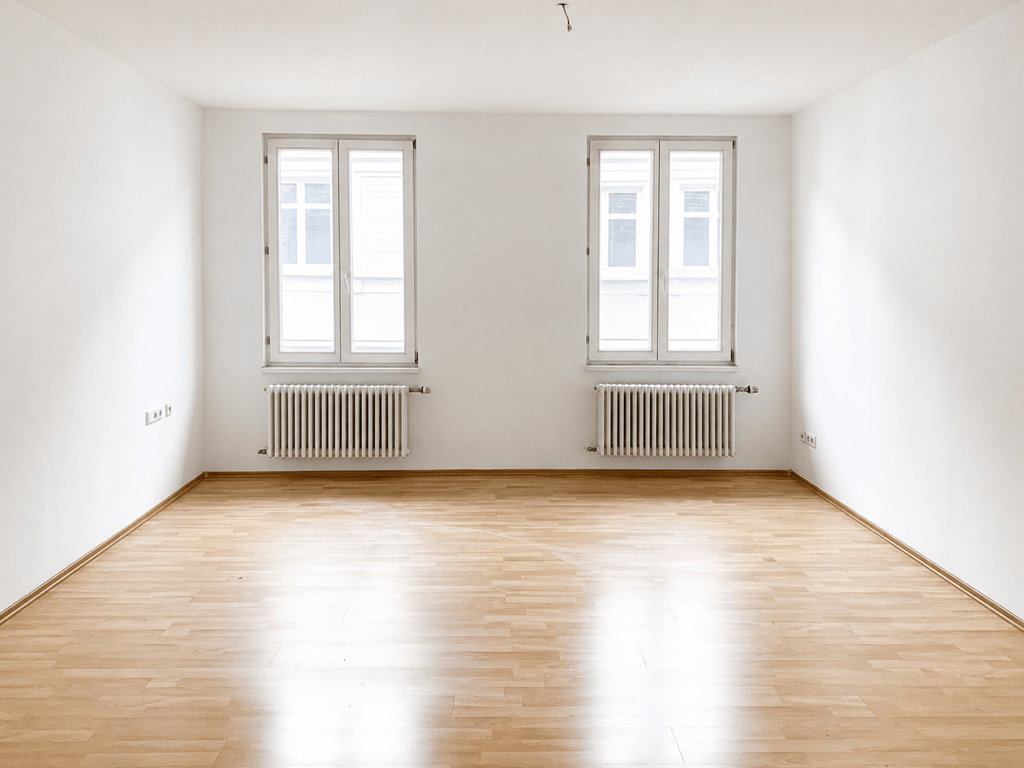 Ein leerer Raum mit weißen Wänden, zwei großen Fenstern, zwei weißen Heizkörpern unter den Fenstern und einem hellen Holzboden.