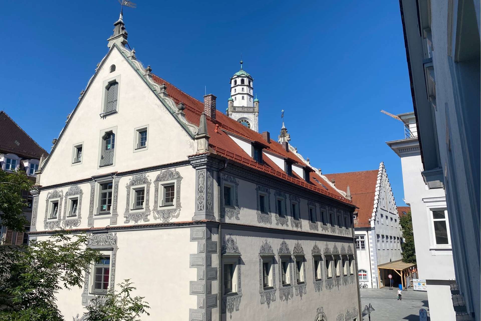 Historisches europäisches Gebäude mit kunstvollen grauen Verzierungen, rotem Ziegeldach und zahlreichen Fenstern, im Hintergrund ein Turm vor strahlend blauem Himmel. Angrenzende Gebäude und Bäume sind teilweise zu sehen.
