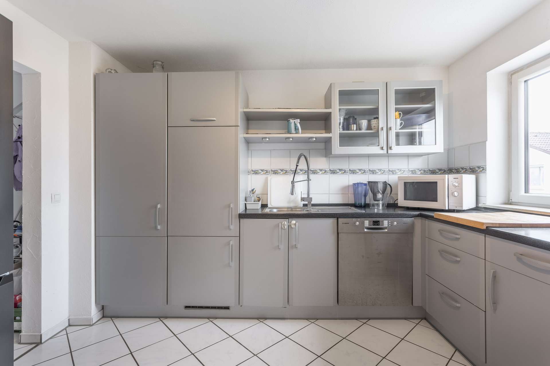 Moderne Küche mit grauen Schränken, Spüle, Geschirrspüler, Mikrowelle und Küchengeräten auf der Arbeitsplatte. Sonnenlicht fällt durch ein Fenster auf der rechten Seite und erhellt den sauberen, aufgeräumten Raum mit Fliesenboden und Schränken mit Glasfronten.