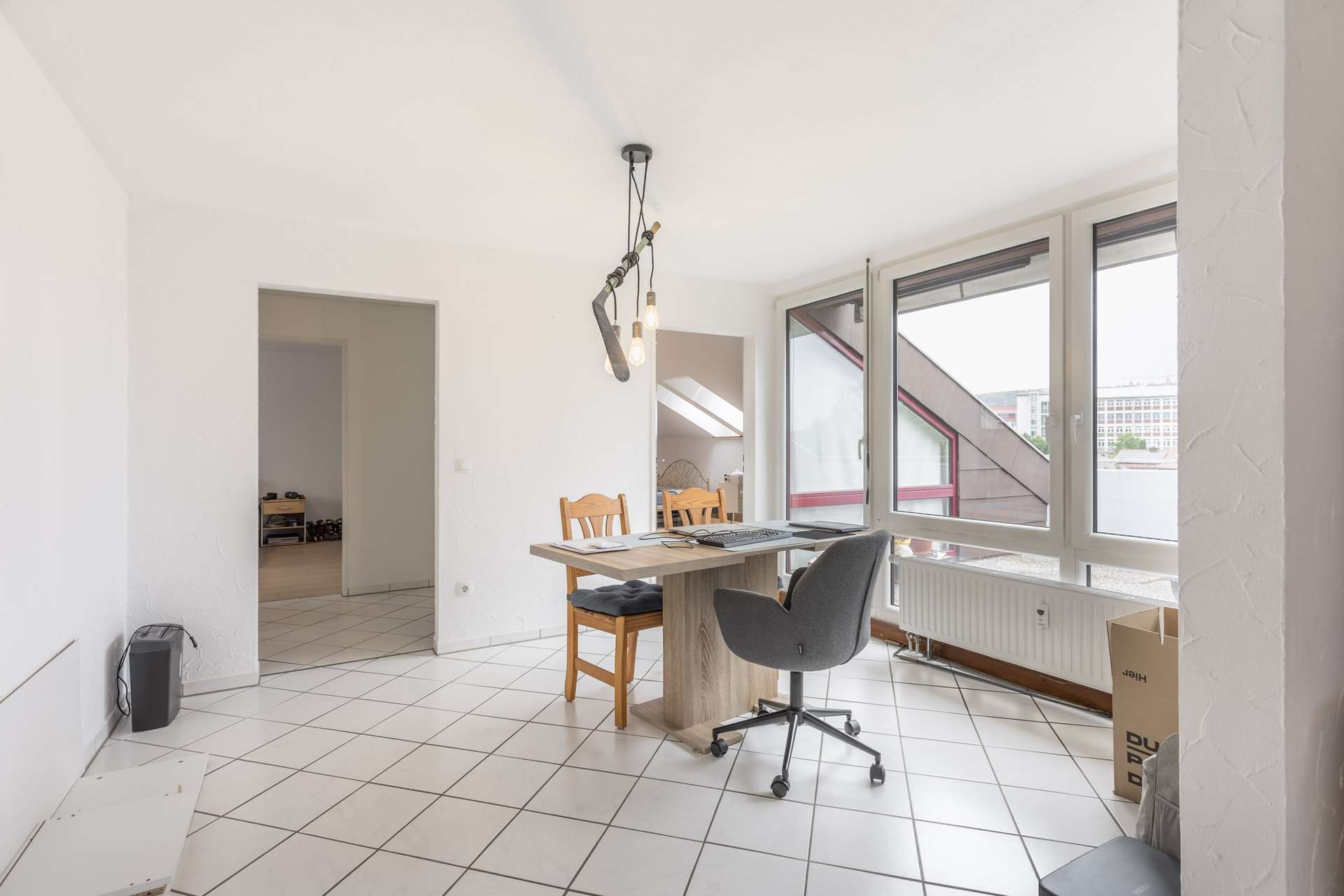 Ein heller Raum mit weißem Fliesenboden, einem kleinen Esstisch mit zwei Holzstühlen und einem grauen Bürostuhl, großen Fenstern und einigen Umzugskartons in der Ecke. Über dem Tisch hängt eine moderne Deckenleuchte.