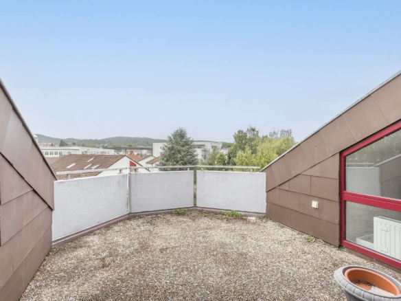 Kleine Dachterrasse mit Kiesboden, Glasgeländer und braunen, schrägen Wänden. Der Blick reicht über Dächer, Bäume und ferne Hügel unter einem strahlend blauen Himmel.
