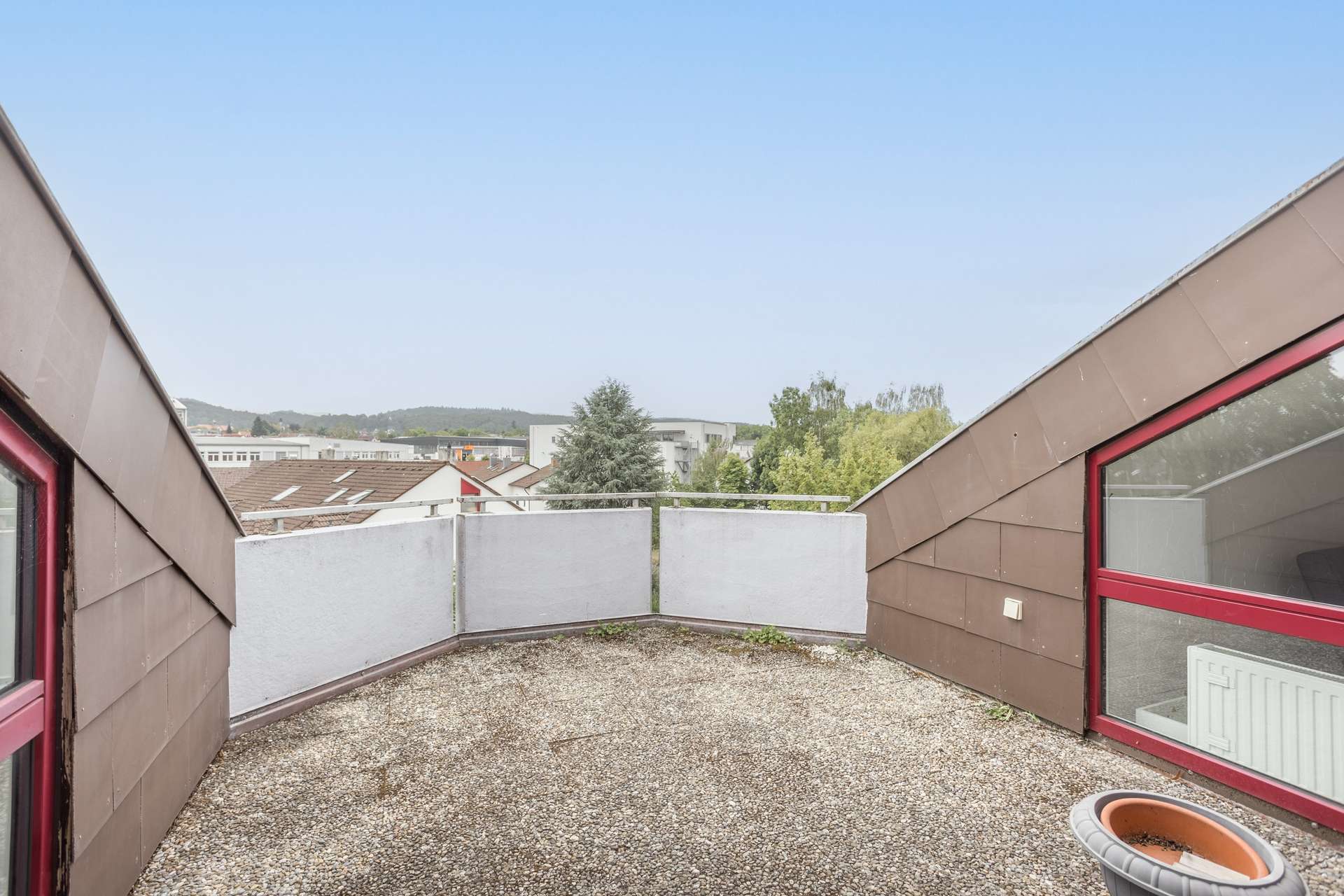 Kleine Dachterrasse mit Kiesboden, Glasgeländer und braunen, schrägen Wänden. Der Blick reicht über Dächer, Bäume und ferne Hügel unter einem strahlend blauen Himmel.
