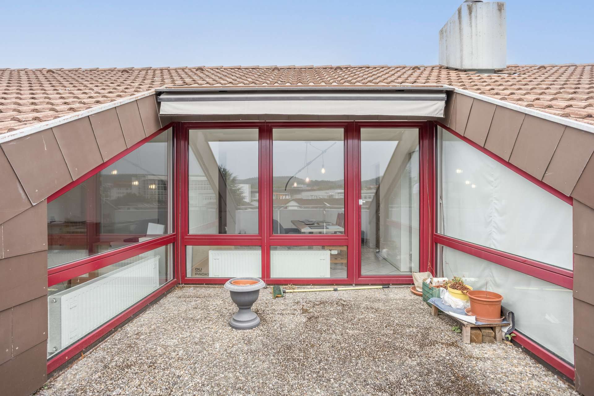 Eine kleine Dachterrasse mit Kiesboden, Topfpflanzen und einem runden Sockel. Große Fenster mit roten Rahmen und Glastüren führen ins Innere des Gebäudes. Im Hintergrund sind Dächer und der Himmel zu sehen.