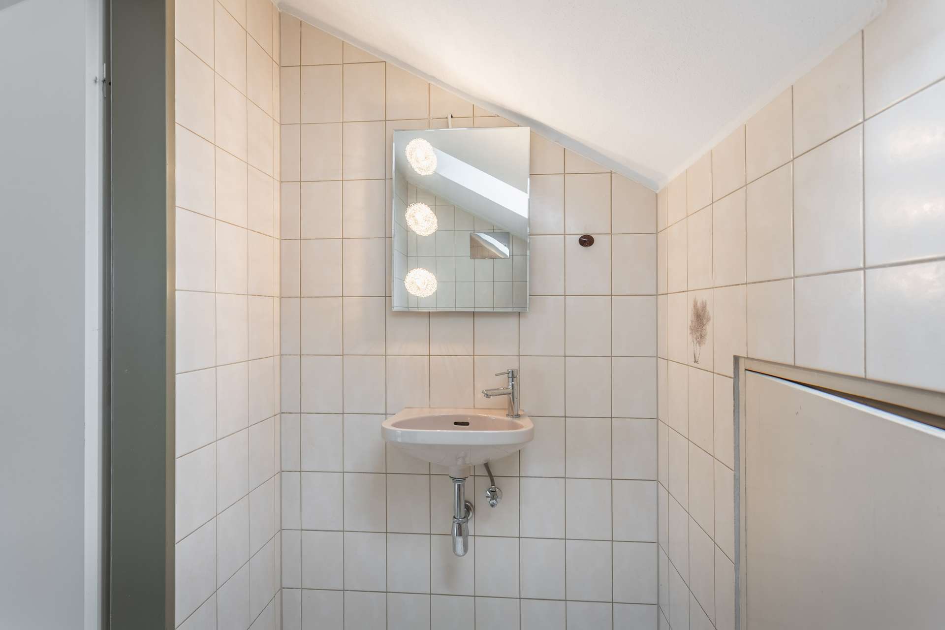 Ein kleines Badezimmer mit beige gefliesten Wänden, einem wandmontierten Waschbecken, einem rechteckigen Spiegel darüber und drei runden Lampen, die sich im Spiegel spiegeln. An der rechten Wand ist ein Fleck zu sehen, und darüber verläuft die Decke schräg.