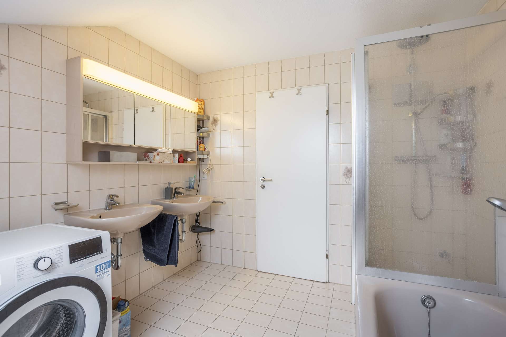 Badezimmer mit beigen Fliesen, zwei Waschbecken mit Spiegeln darüber, einer Waschmaschine, einer Duschkabine aus Milchglas und Handtuchhaltern. An der Wand befinden sich Regale mit Toilettenartikeln, und in der Mitte ist eine weiße Tür zu sehen.
