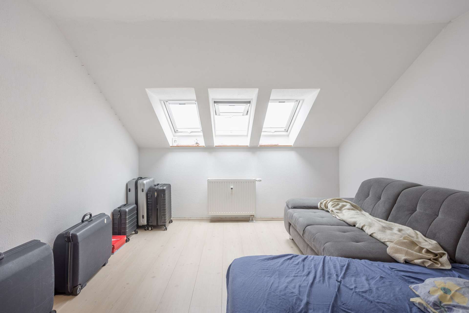 Minimalistisch eingerichtetes Dachzimmer mit hellem Holzboden, drei Dachfenstern, mehreren Koffern an einer Wand, einem Heizkörper unter den Fenstern, einem grauen Sofa mit einer beigen Decke und einem Teil eines mit blauem Stoff bezogenen Bettes im Vordergrund.