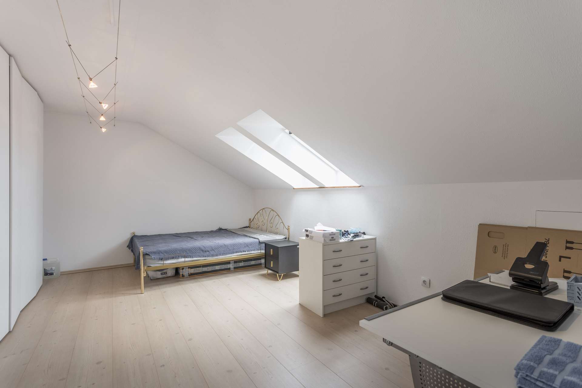Minimalistisch eingerichtetes Schlafzimmer im Dachgeschoss mit einem Einzelbett, Dachfenstern, einer kleinen Kommode und einem Schreibtisch mit Büroartikeln. Weiße Wände, ein heller Holzboden und moderne Beleuchtung schaffen einen hellen, aufgeräumten Raum.