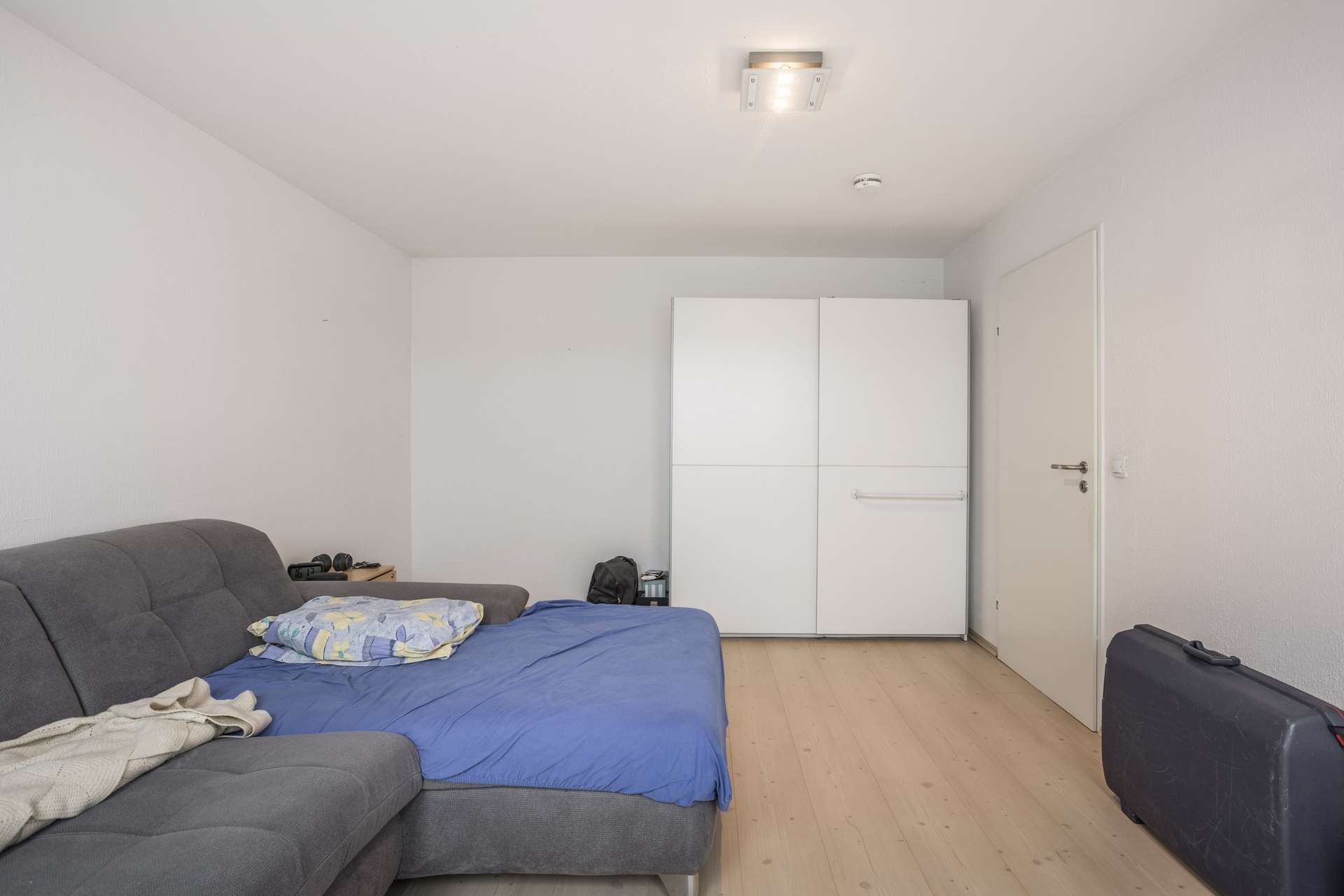 Ein schlichter Raum mit einem grauen Schlafsofa, das mit einer blauen Decke bedeckt ist, einem weißen Kleiderschrank mit geschlossener Tür und einem schwarzen Koffer auf einem hellen Holzboden. Auf dem Sofa liegen ein Kissen und einige Kleidungsstücke.