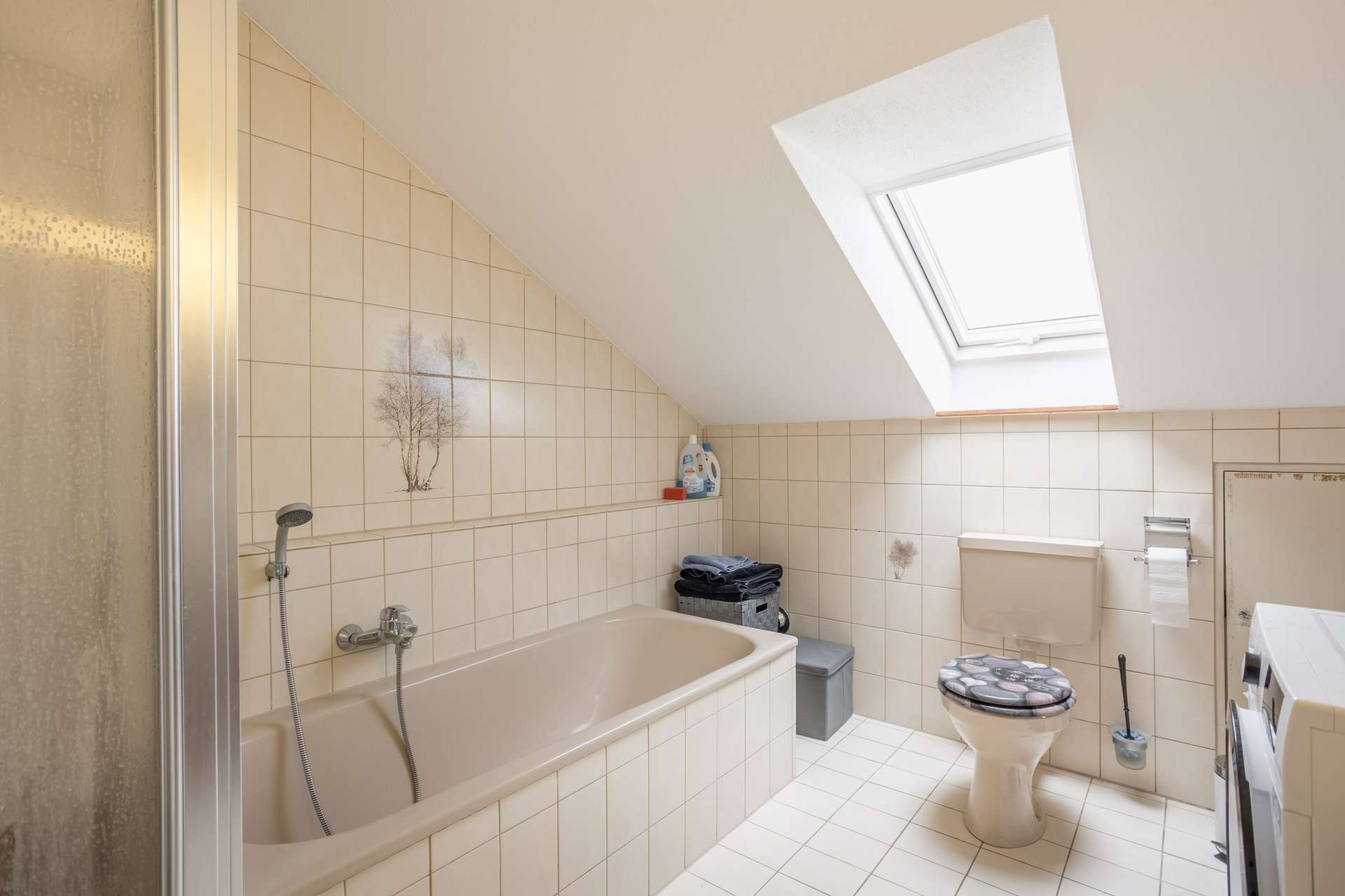 Badezimmer mit beigen Fliesen, einer Badewanne, einer Toilette, einer Waschmaschine und einer schrägen Decke mit einem Dachfenster. Außerdem sind eine Dusche und Reinigungsmittel zu sehen. Der Raum ist sauber und wird von Tageslicht gut beleuchtet.