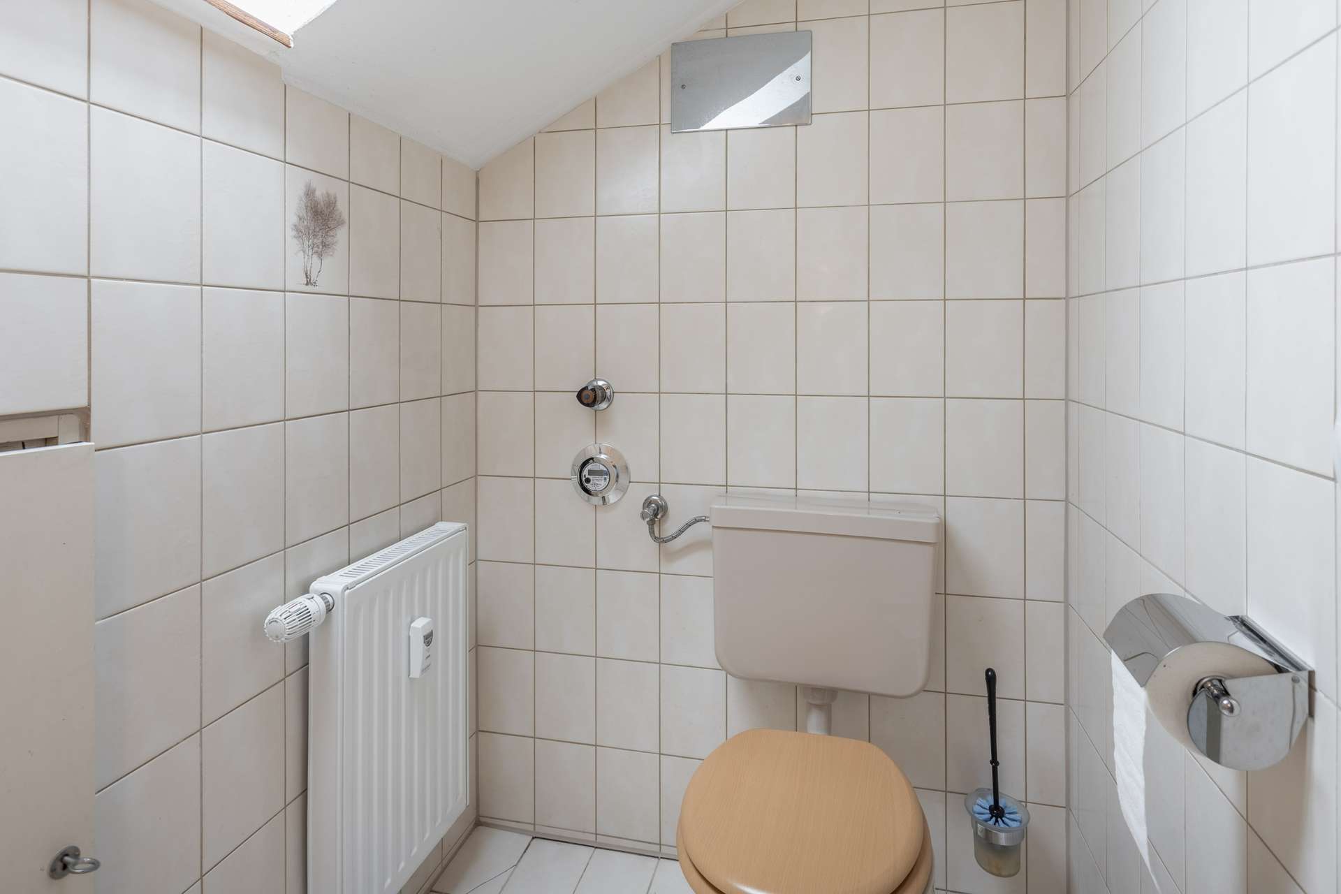 Kleines Badezimmer mit beige gefliesten Wänden, einer Toilette mit beigem Sitz, einem wandmontierten Spülkasten, einem Heizkörper, einem Toilettenpapierhalter und einer Toilettenbürste. Auf einer Fliese befindet sich ein kleines Pflanzenmotiv als Aufkleber.