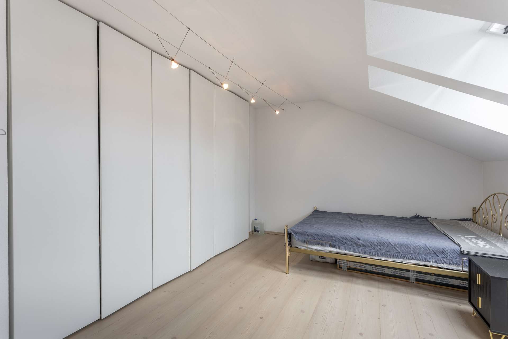 Ein minimalistisches Schlafzimmer mit schräger Decke, großen weißen Kleiderschränken entlang einer Wand, einem Bett mit grauer Bettwäsche, hellem Holzboden und Oberlichtern, die für Tageslicht sorgen.