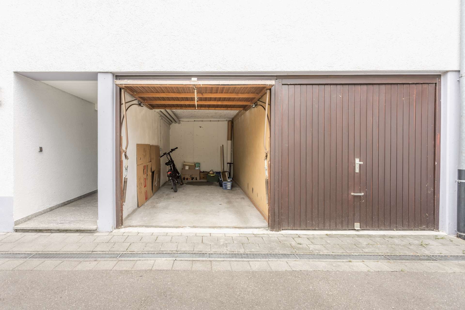 Eine Einzelgarage mit einem teilweise geöffneten Holztor, in der ein Fahrrad, Kisten und verschiedene Werkzeuge stehen. Die Garage ist an ein weißes Gebäude angebaut, vor dem sich eine gepflasterte Einfahrt befindet.