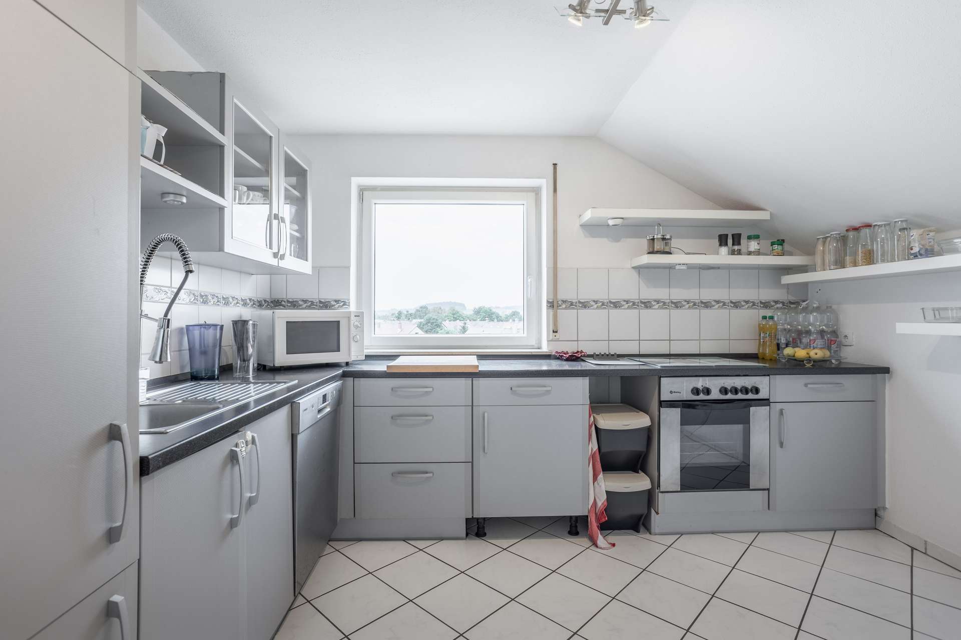 Moderne Küche mit grauen Schränken, einer Spüle, einer Mikrowelle, einem Herd und offenen Regalen. Ein Fenster über der Arbeitsplatte lässt Tageslicht herein. Der Boden und die Fliesenwand sind weiß gefliest. In den Regalen stehen Kochutensilien und Gläser.
