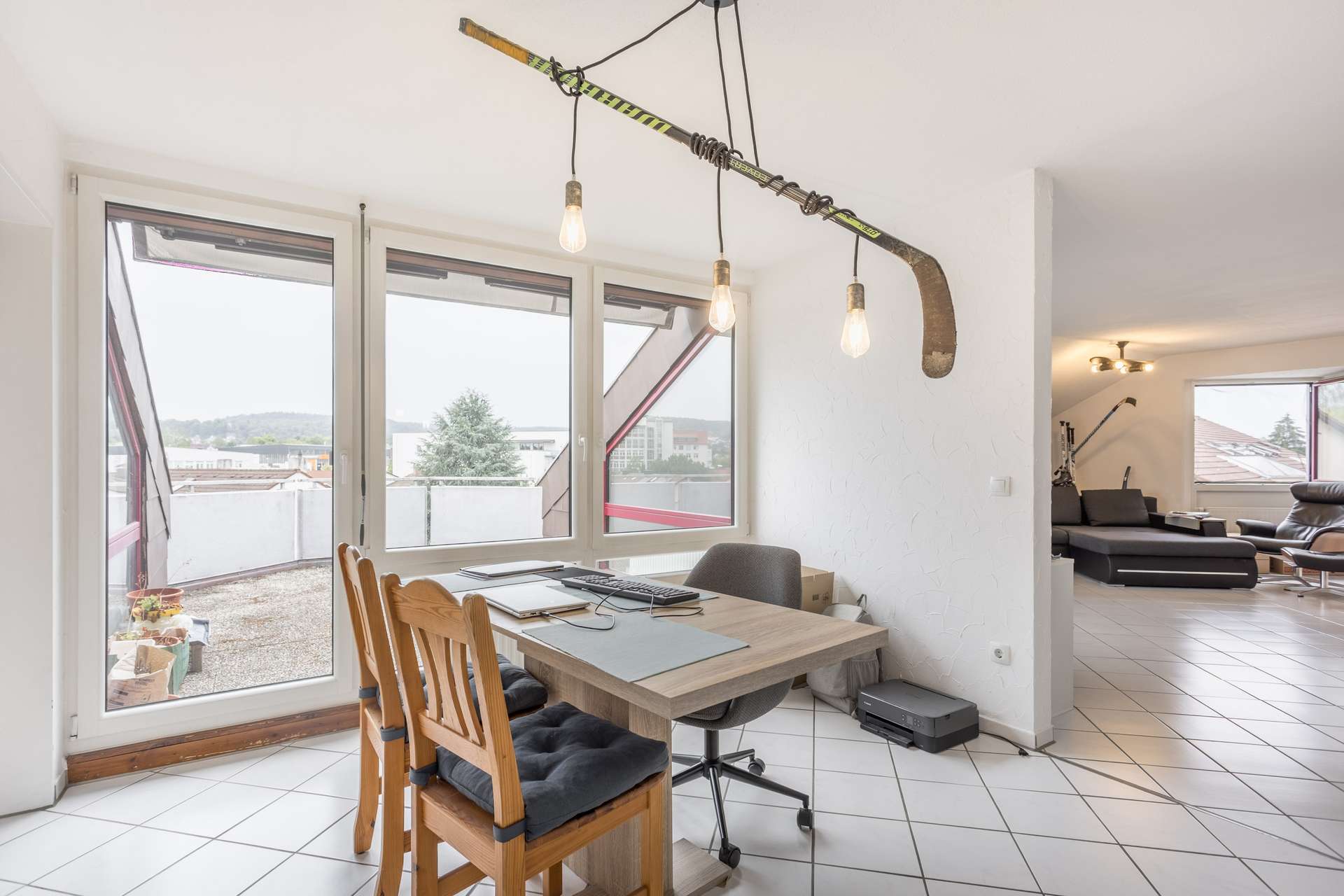 Ein helles Büro mit großen Fenstern, einem Schreibtisch und Stühlen sowie einer Deckenlampe aus einem Hockeyschläger. Der Raum geht in einen modernen Wohnbereich mit schwarzen Sofas und weiß gefliesten Böden über.