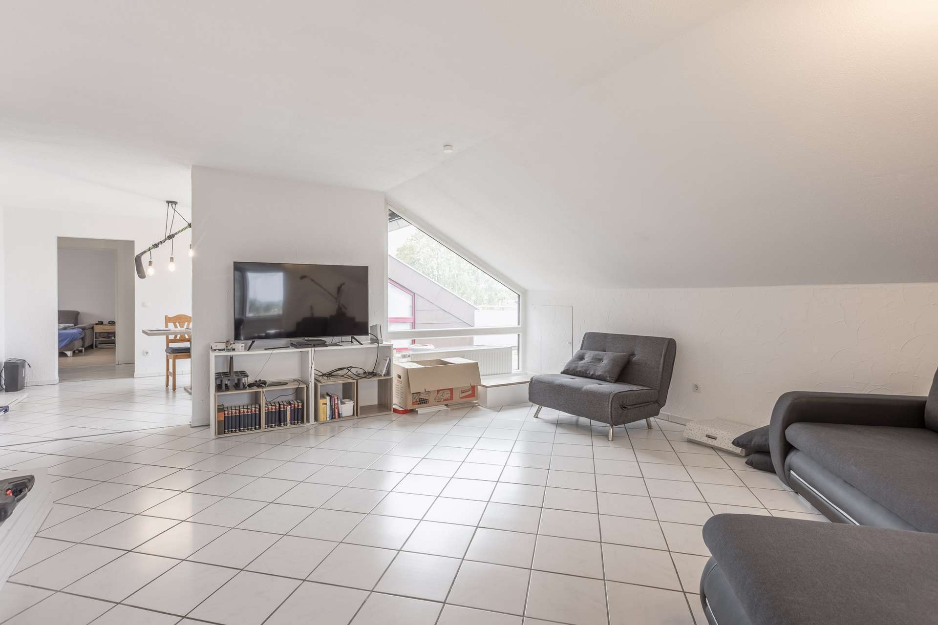 Helles Wohnzimmer mit weißem Fliesenboden, grauen Sofas, einem Fernseher auf einem Ständer, Büchern und einem großen Fenster. Im Hintergrund sind der Essbereich und das Schlafzimmer zu sehen. Neben dem Fenster stehen Kartons.