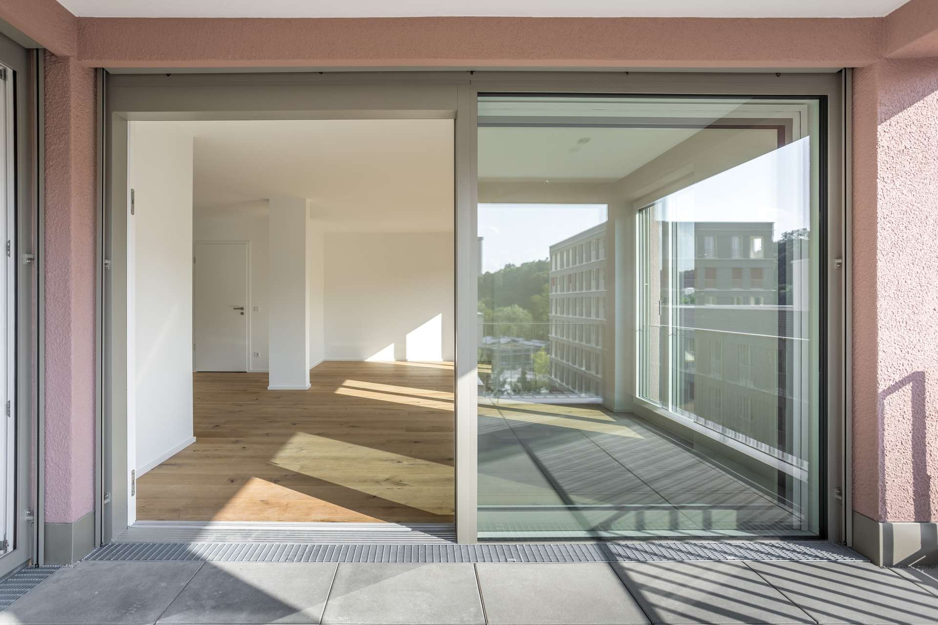Loggia Der Blick durch eine große Glasschiebetür fällt in einen hellen, leeren Raum mit Holzböden und weißen Wänden; draußen sind Gebäude und Grünflächen zu sehen.