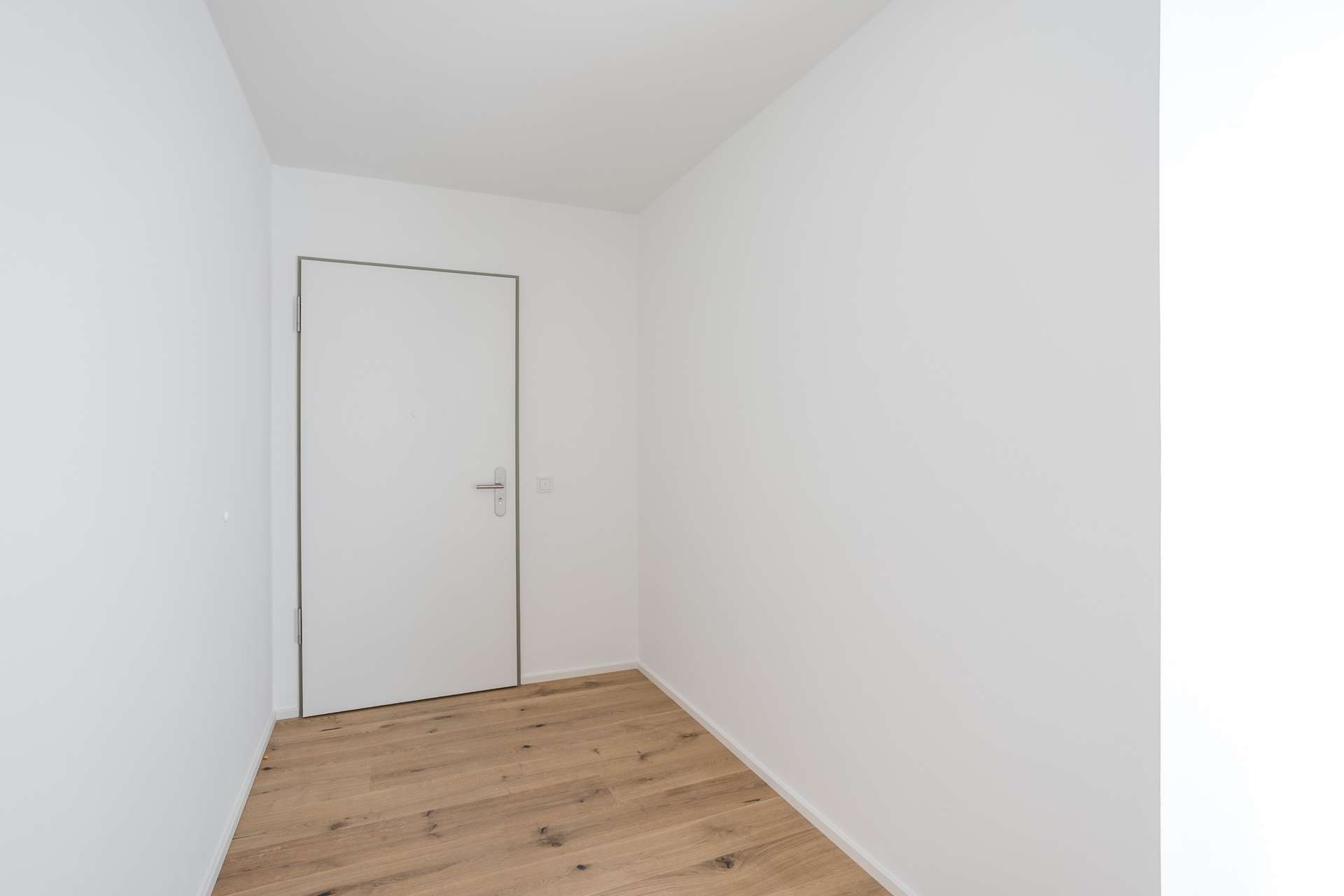 Eingang Ein kleiner, leerer weißer Raum mit einer geschlossenen weißen Tür und hellem Holzfußboden. Der Raum ist hell und minimalistisch eingerichtet; es sind weder Möbel noch Dekorationen zu sehen.