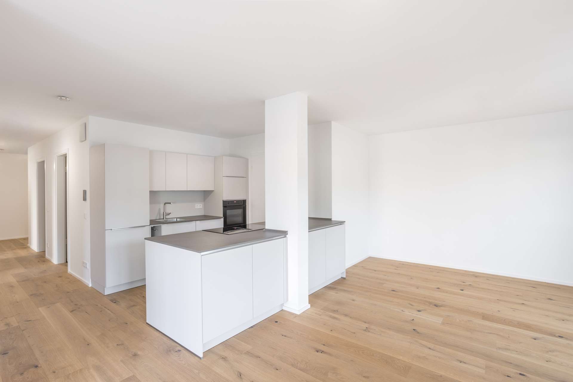 Wohn- und Essbereich Moderne, minimalistische Küche mit weißen Schränken und grauen Arbeitsplatten, die zu einem großzügigen Raum mit hellen Holzböden und weißen Wänden hin offen ist. Der Raum ist hell und von Tageslicht durchflutet, was ihm eine klare, luftige Atmosphäre verleiht.