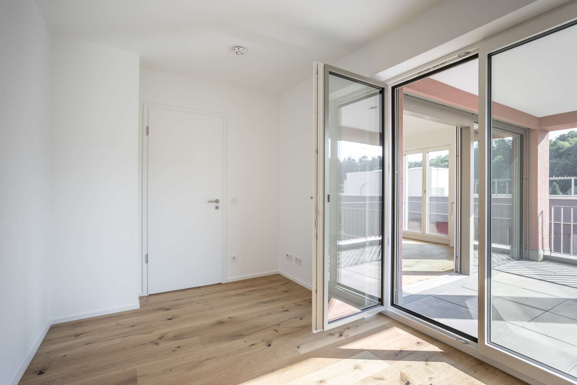 Schlafen 1 Ein heller, leerer Raum mit Holzboden, weißen Wänden, einer offenen Tür, die auf einen Balkon führt, und großen Glasfenstern, durch die Tageslicht hereinfällt.