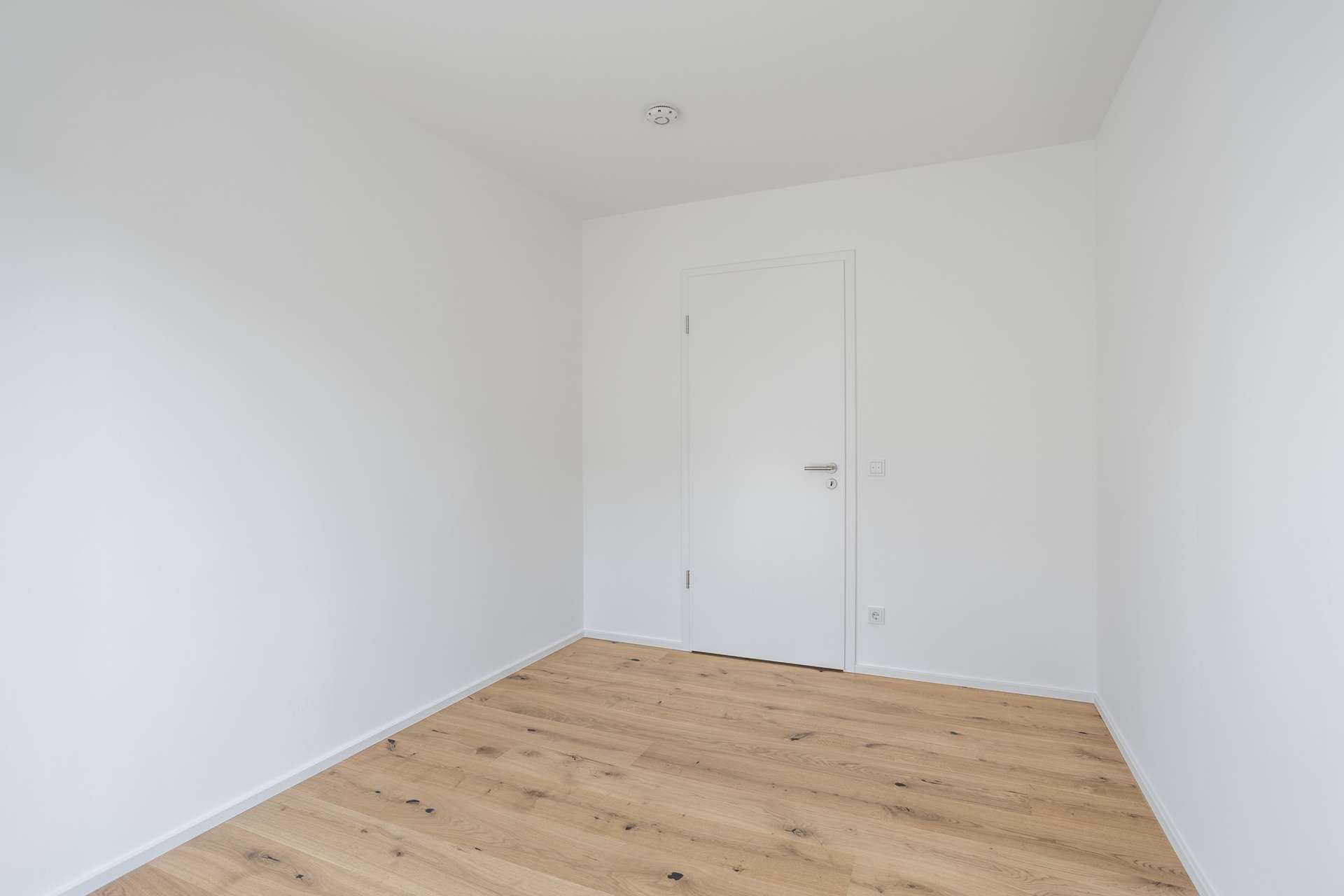 Schlafen 3 Ein kleiner, leerer Raum mit weißen Wänden, einer geschlossenen weißen Tür, einer Deckenlampe und hellem Holzfußboden. Der Raum ist schlicht und minimalistisch gestaltet.