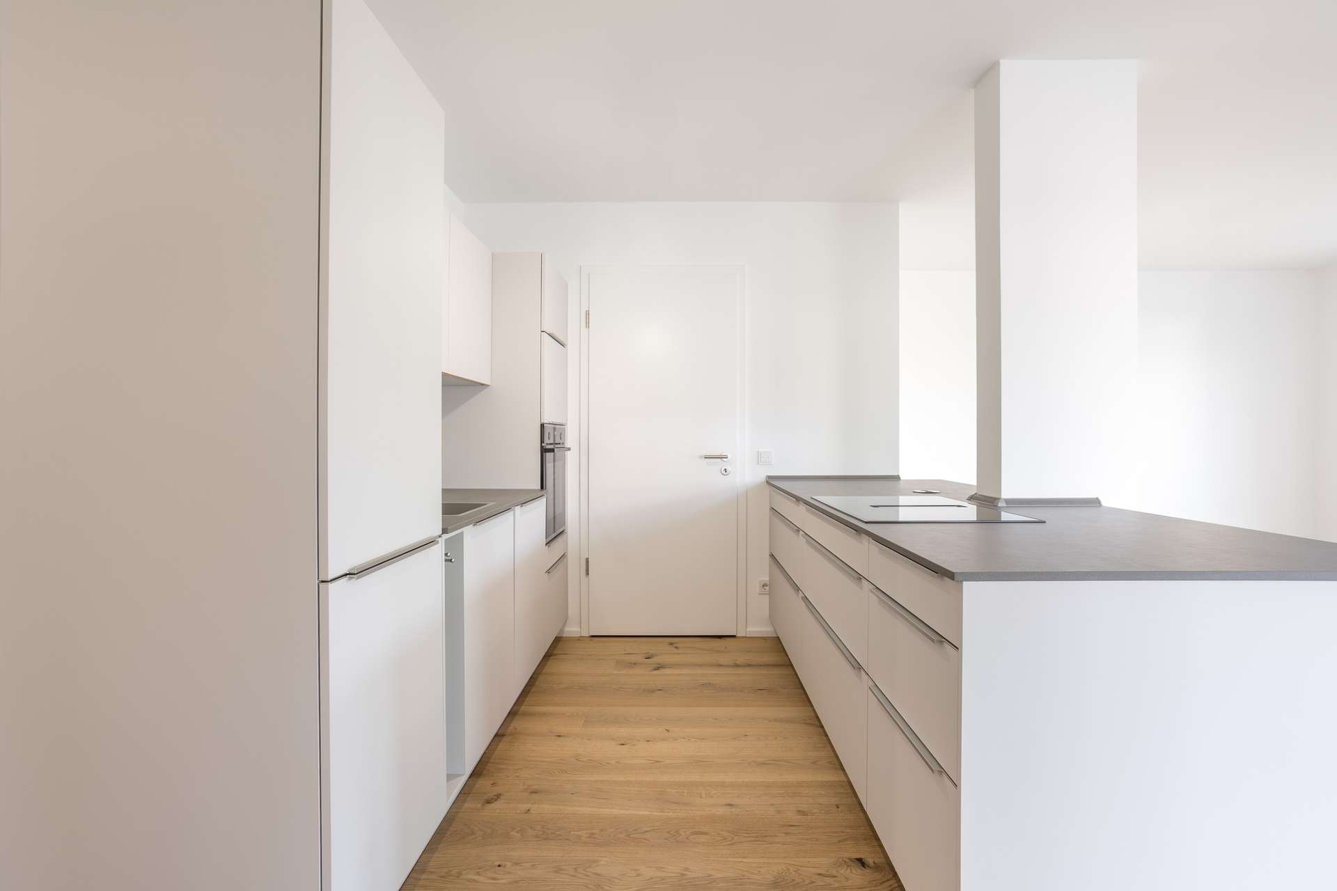 Wohn- und Essbereich Minimalistische, moderne Küche mit weißen Schränken, grauen Arbeitsplatten, Einbaugeräten und hellem Holzboden. Der Raum ist hell, besticht durch klare Linien und eine neutrale Farbpalette.