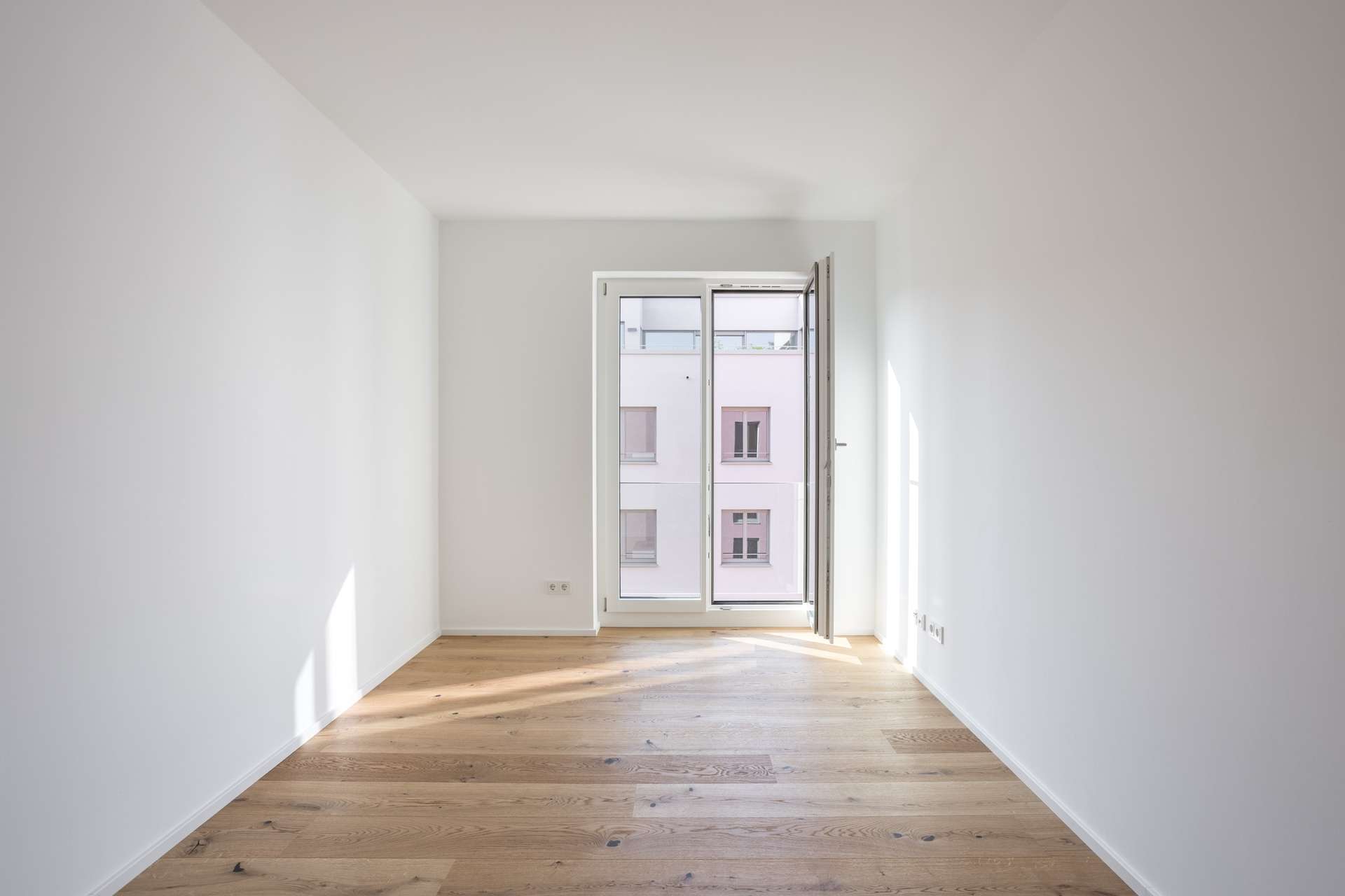 Schlafen 3 Ein kleiner, leerer Raum mit weißen Wänden, Holzböden und einer offenen Glastür, die auf einen Balkon führt. Sonnenlicht fällt durch die Tür und wirft Schatten auf den Boden. Draußen ist ein Wohnhaus zu sehen.