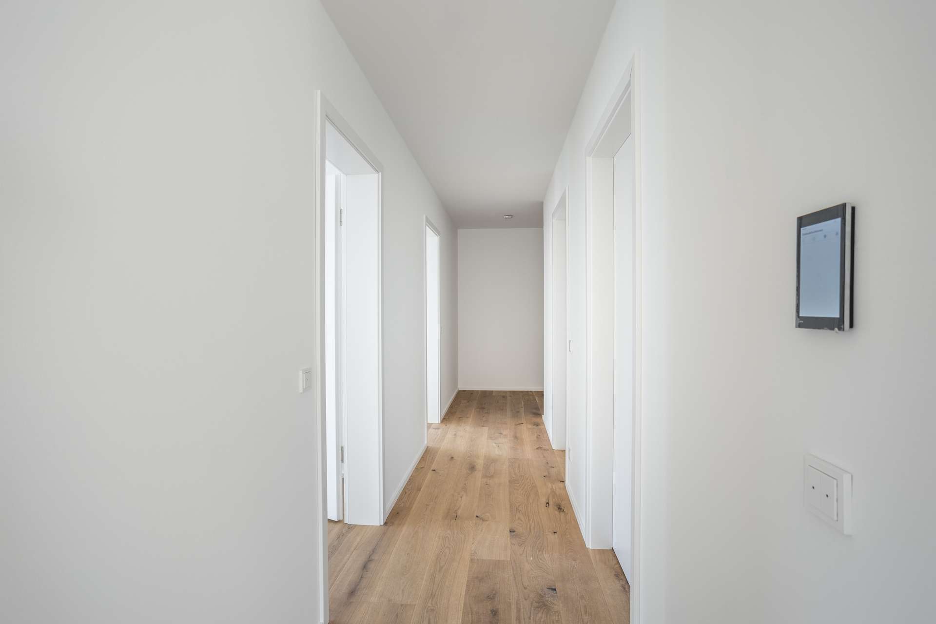 Flur Ein heller, minimalistisch gestalteter Flur mit weißen Wänden, mehreren Türen und hellem Holzboden. An der rechten Wand ist in der Nähe eines Lichtschalters ein digitales Thermostat angebracht.