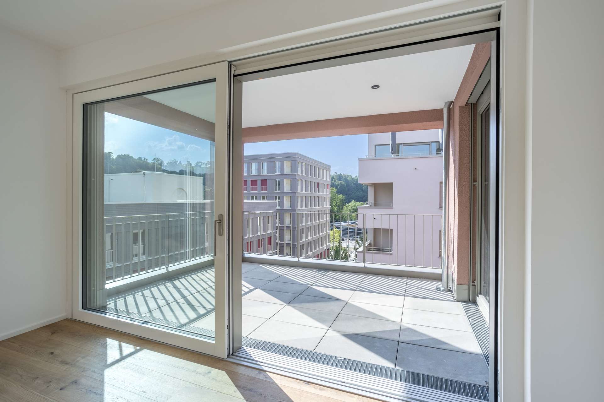 Loggia Eine moderne Wohnungseinrichtung mit großen Glasschiebetüren, die auf einen großzügigen Balkon führen. Draußen sind benachbarte Wohnhäuser und Grünflächen zu sehen. Sonnenlicht erhellt den Holzboden und den gefliesten Balkon.