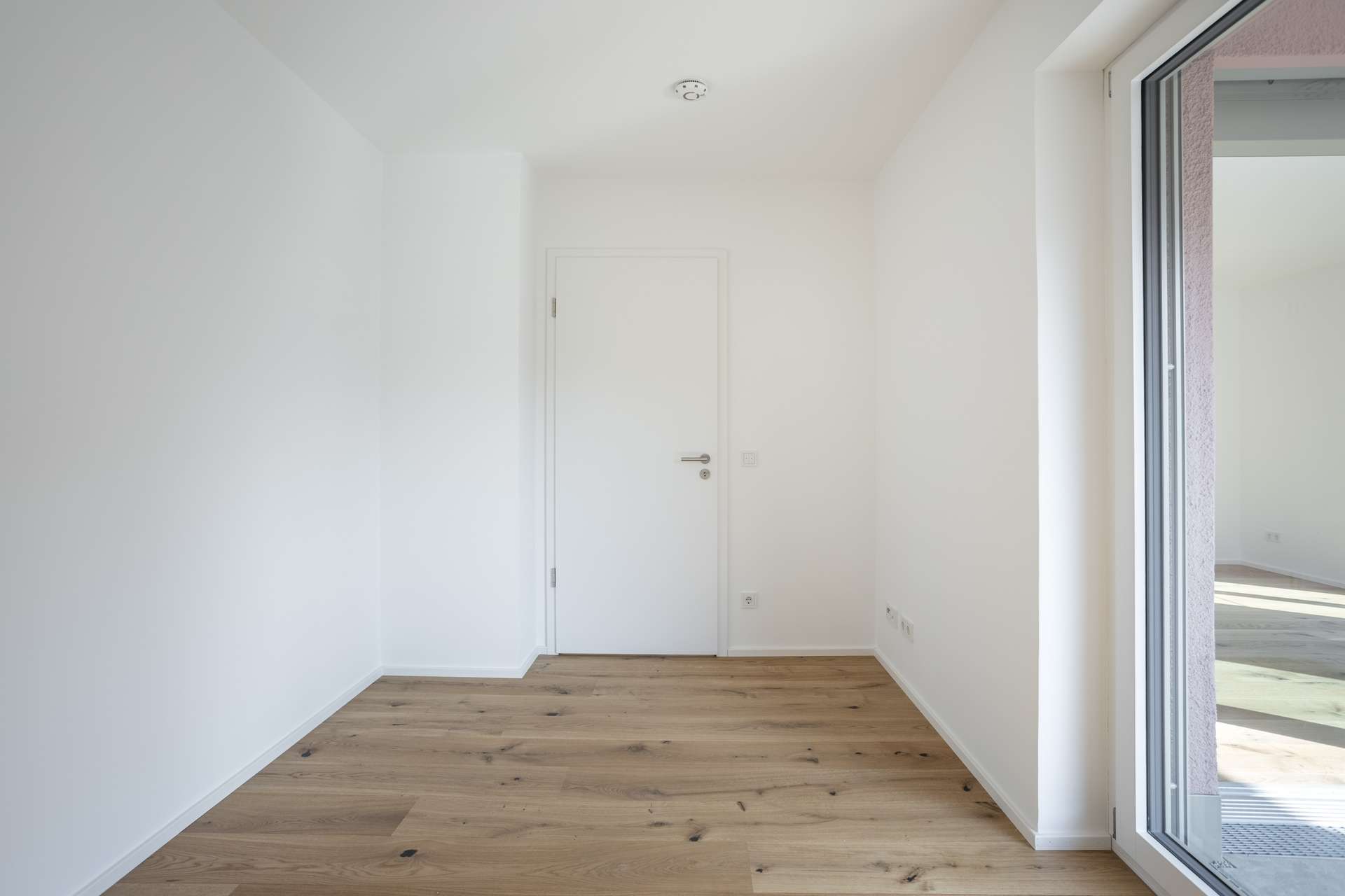 Schlafen 1 Ein kleiner, leerer Raum mit weißen Wänden, einem Holzboden, einer geschlossenen weißen Tür und einer großen Glasschiebetür, durch die Tageslicht hereinfällt.