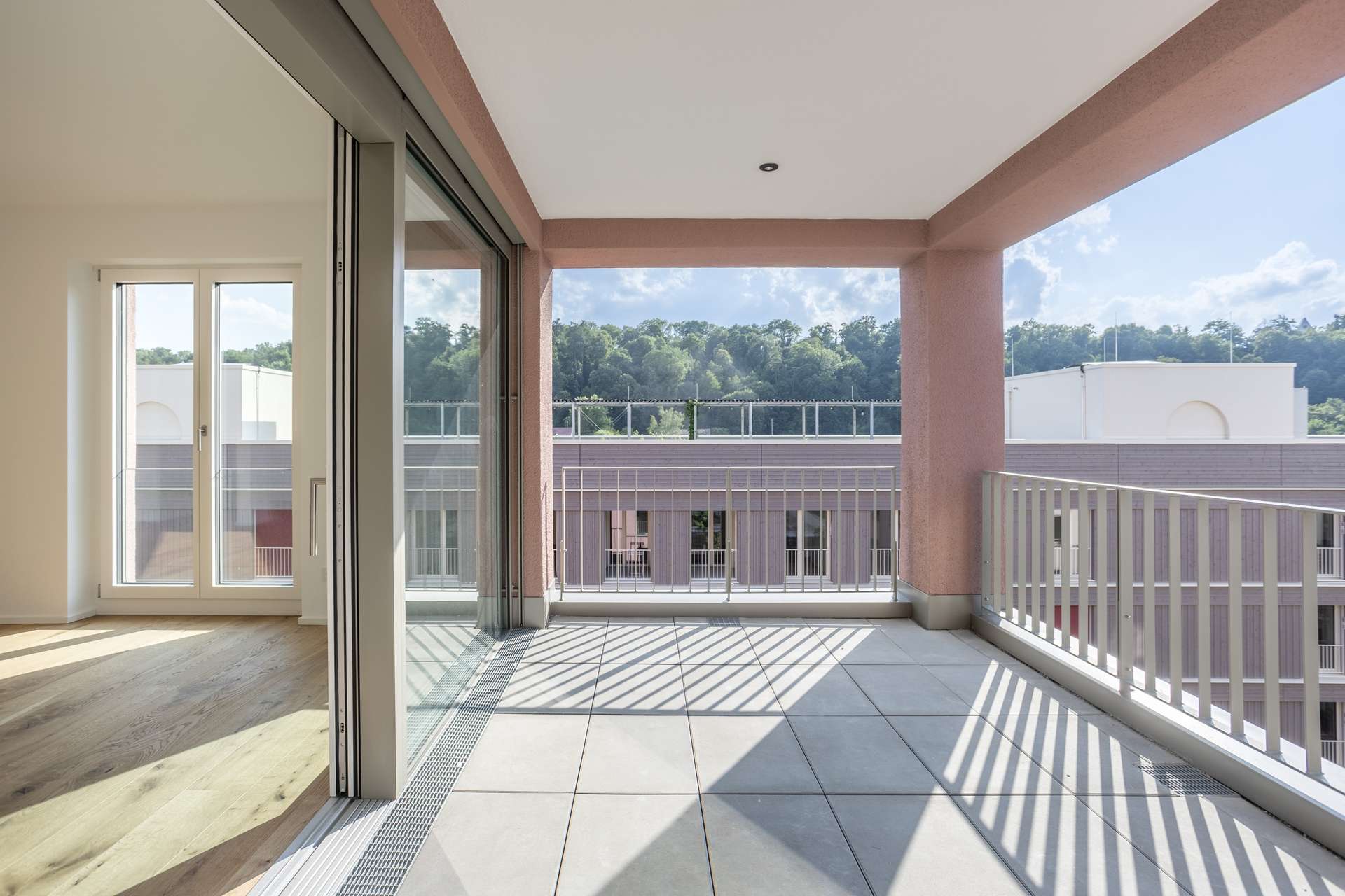 Loggia Ein moderner Wohnungsbalkon mit großen Fliesen, Glasgeländern und Blick auf Bäume und Dächer. Sonnenlicht wirft Schatten auf den Boden, und Glasschiebetüren verbinden den Balkon mit dem Innenraum.