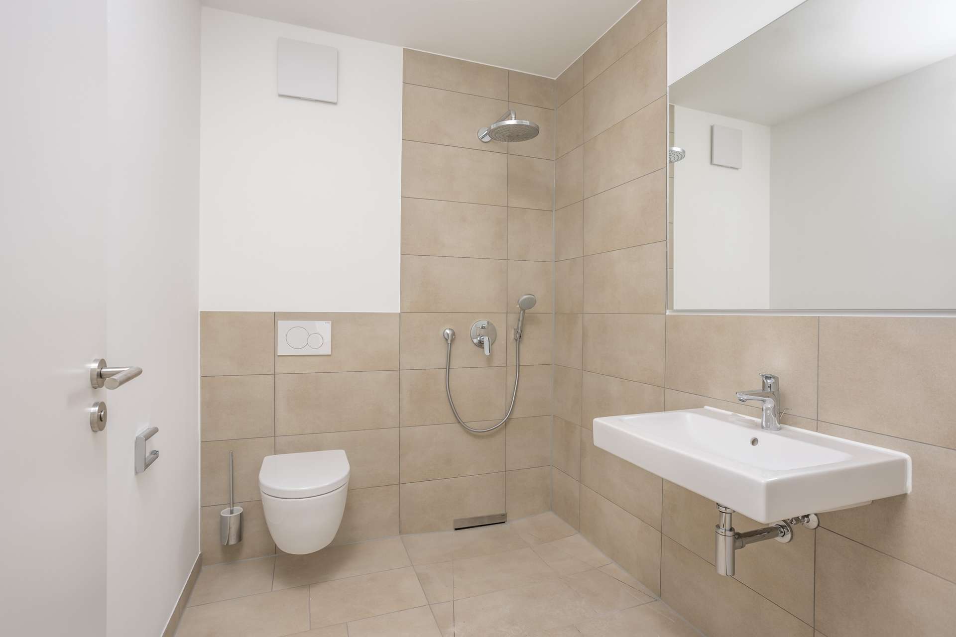 Badezimmer 2 Ein modernes, barrierefreies Badezimmer mit einem wandhängenden WC, einer ebenerdigen Dusche mit Regenduschkopf und Haltegriff, einem weißen Waschbecken, einem großen Spiegel sowie beigen Fliesen an Wänden und Boden.