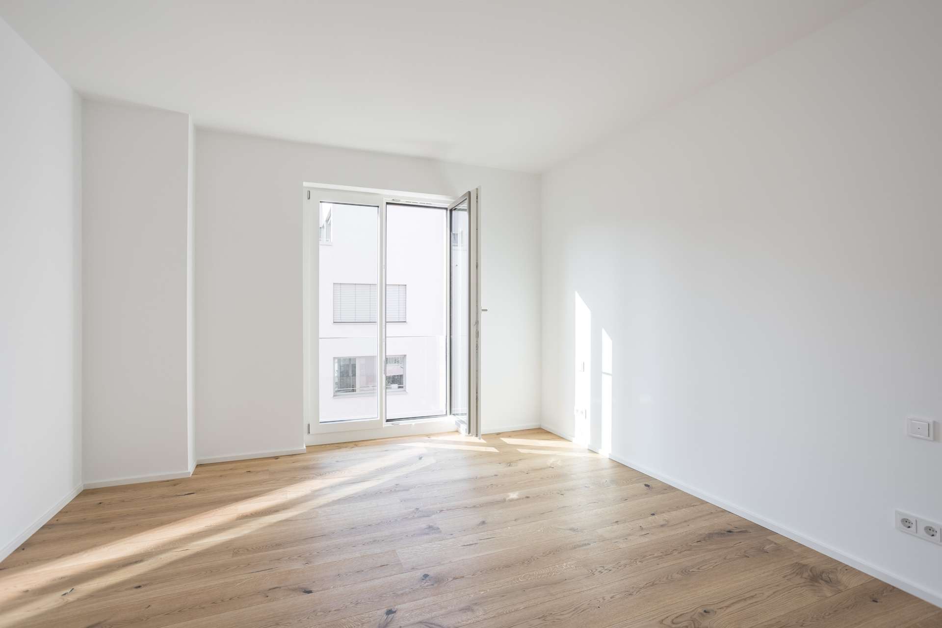 Schlafen 2 Ein heller, leerer Raum mit weißen Wänden, hellem Holzboden und einem großen Fenster, bei dem eine Scheibe geöffnet ist, sodass Tageslicht hereinfällt. Das Fenster bietet einen Blick auf die Fassade eines Gebäudes.