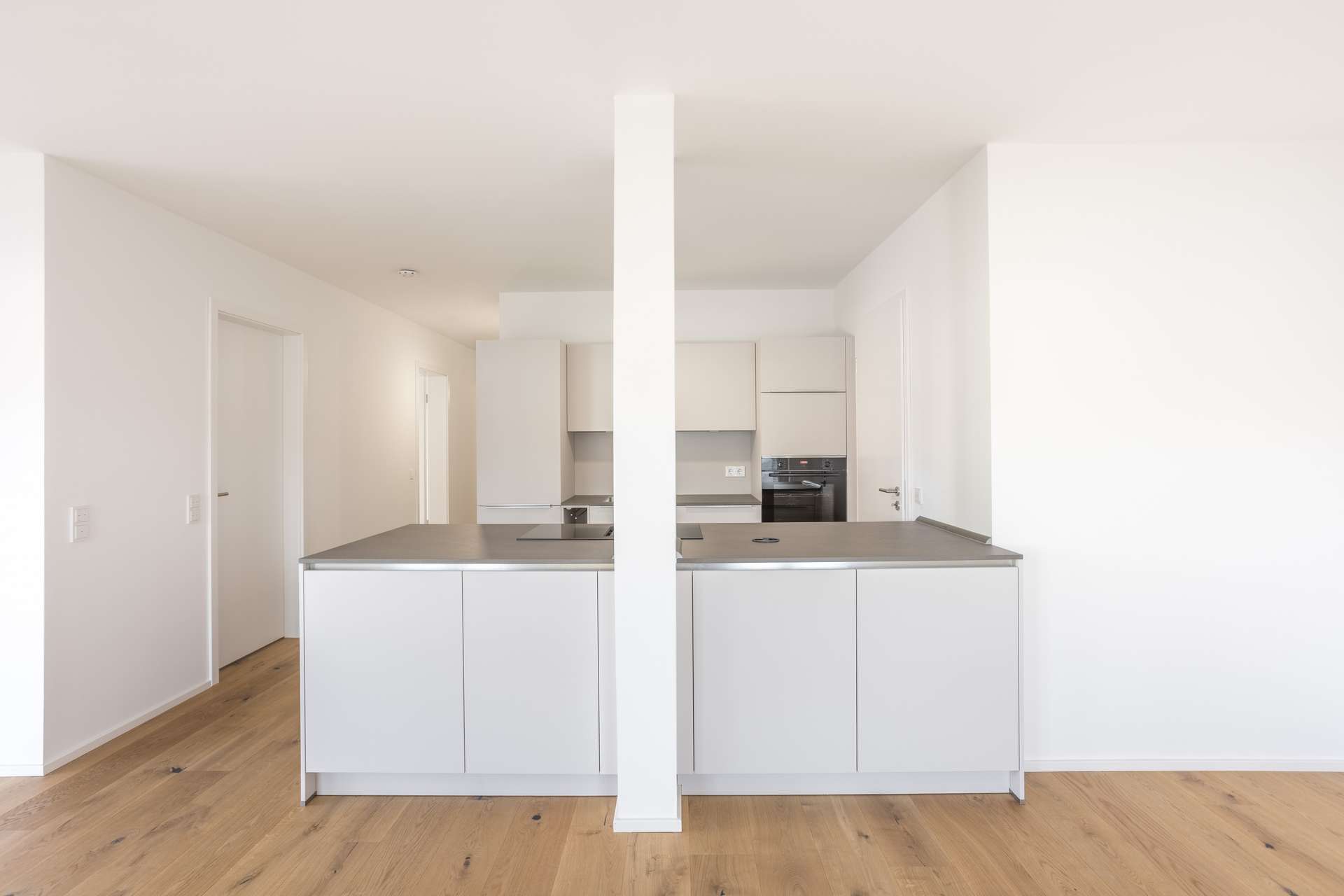 Wohn- und Essbereich Eine moderne, minimalistische Küche mit einer zentralen weißen Trennwand, hellen Holzböden, weißen Schränken und Arbeitsplatten aus Edelstahl, die ein symmetrisches und klares Erscheinungsbild schaffen. Der Raum ist hell und übersichtlich.