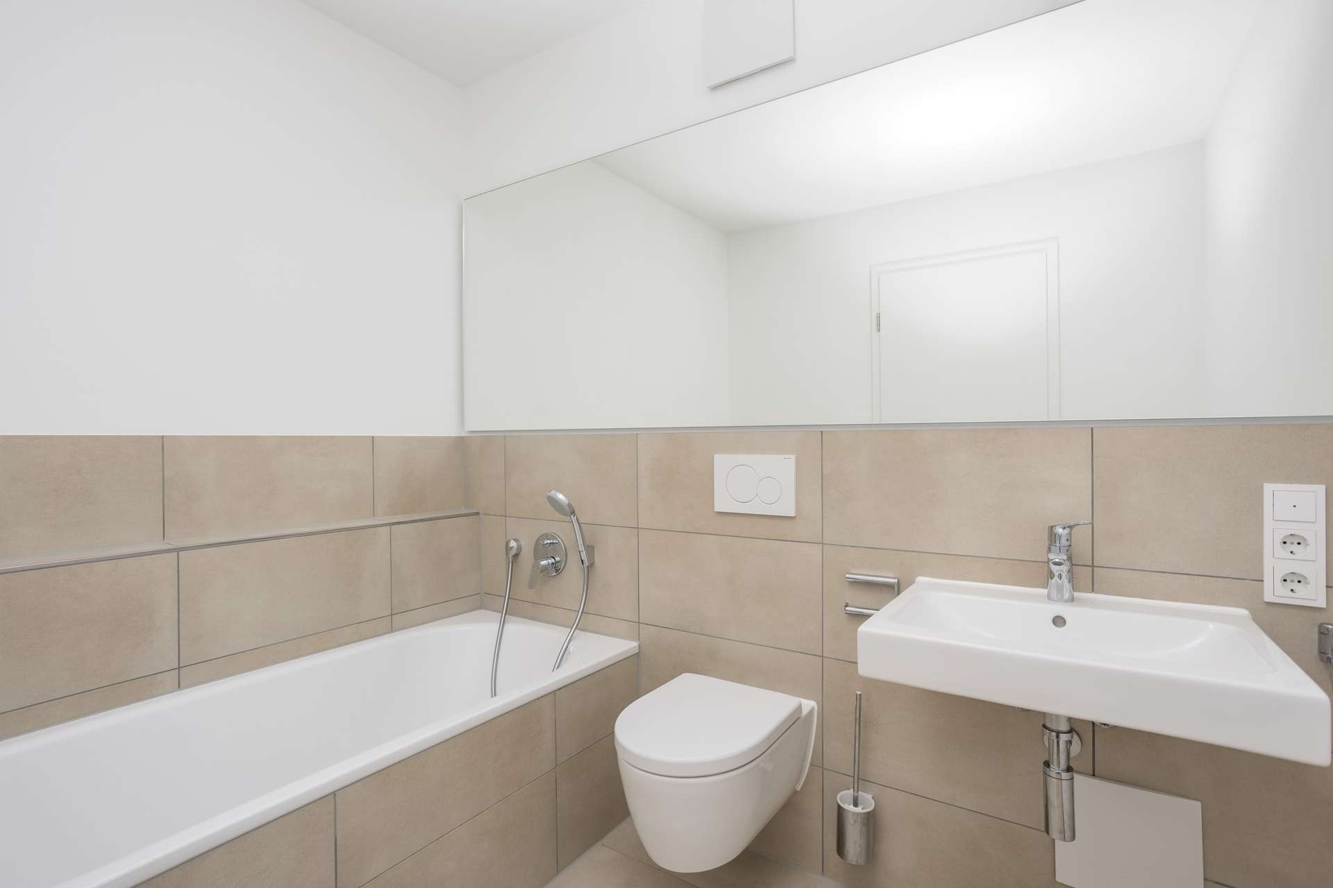 Badezimmer 1 Modernes Badezimmer mit beigen Fliesen, Badewanne mit Duschkopf, wandhängendem WC, weißem Waschbecken, großem Spiegel darüber und weißen Wänden. Minimalistisches und klares Design mit schlichten Armaturen.