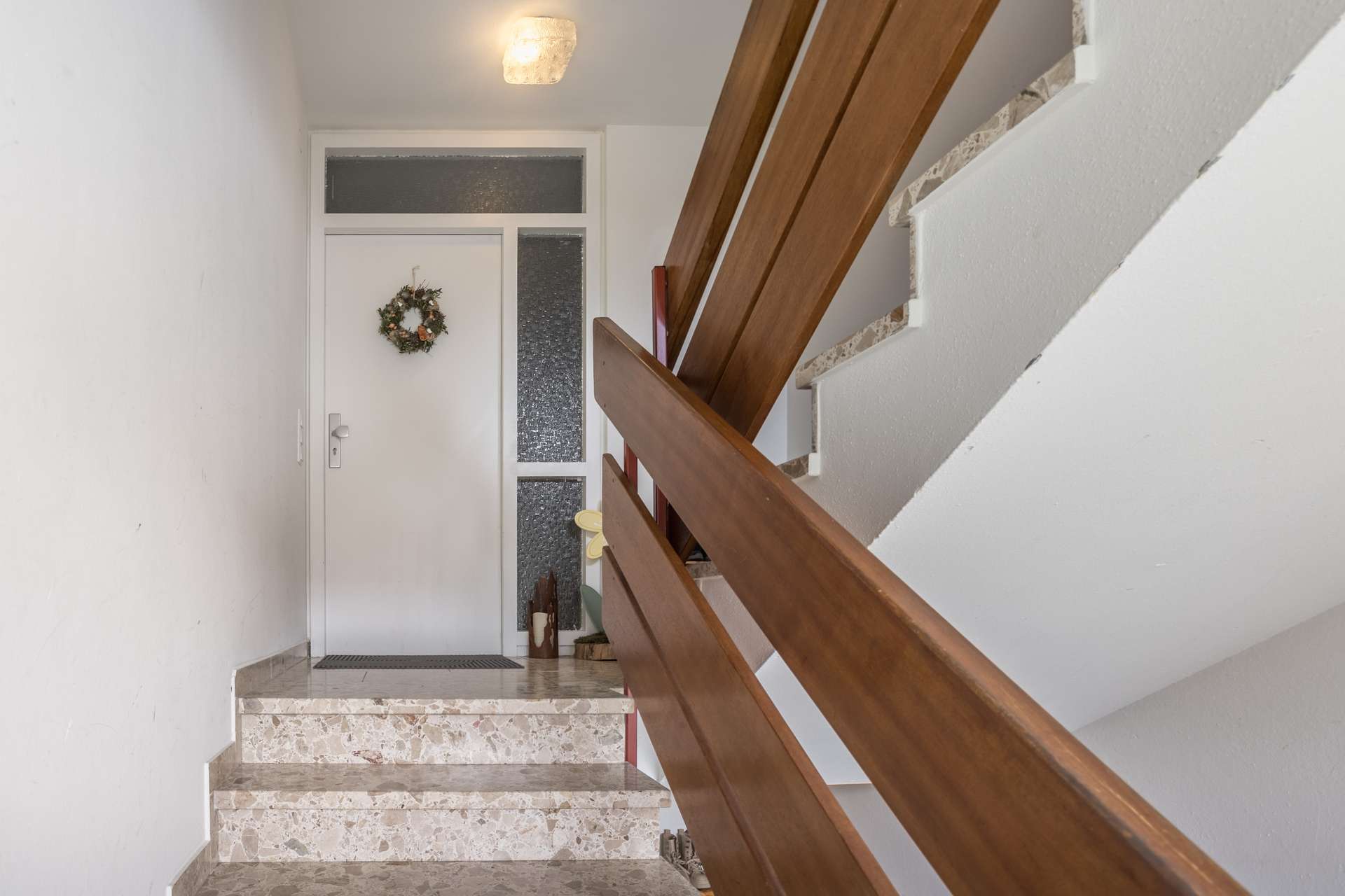 1.OG Blick von einer Steintreppe auf eine weiße Haustür mit einem kleinen Kranz und einem Holzgeländer.