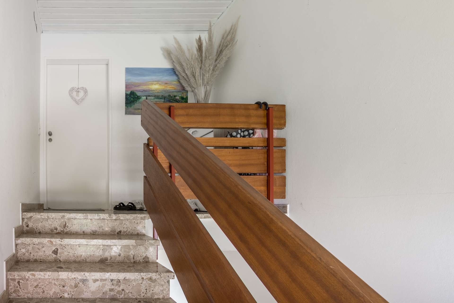 DG Eine Holztreppe mit einem langen Geländer, die zu einem Treppenabsatz führt, auf dem sich eine weiße Tür, ein Gemälde mit einem Sonnenuntergang und eine Vase mit Pampasgras befinden.