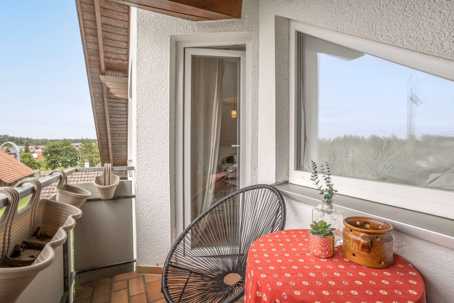 DG Ein Balkon mit einem rot gemusterten Tisch, einem schwarzen Drahtstuhl und ein paar kleinen Topfpflanzen.