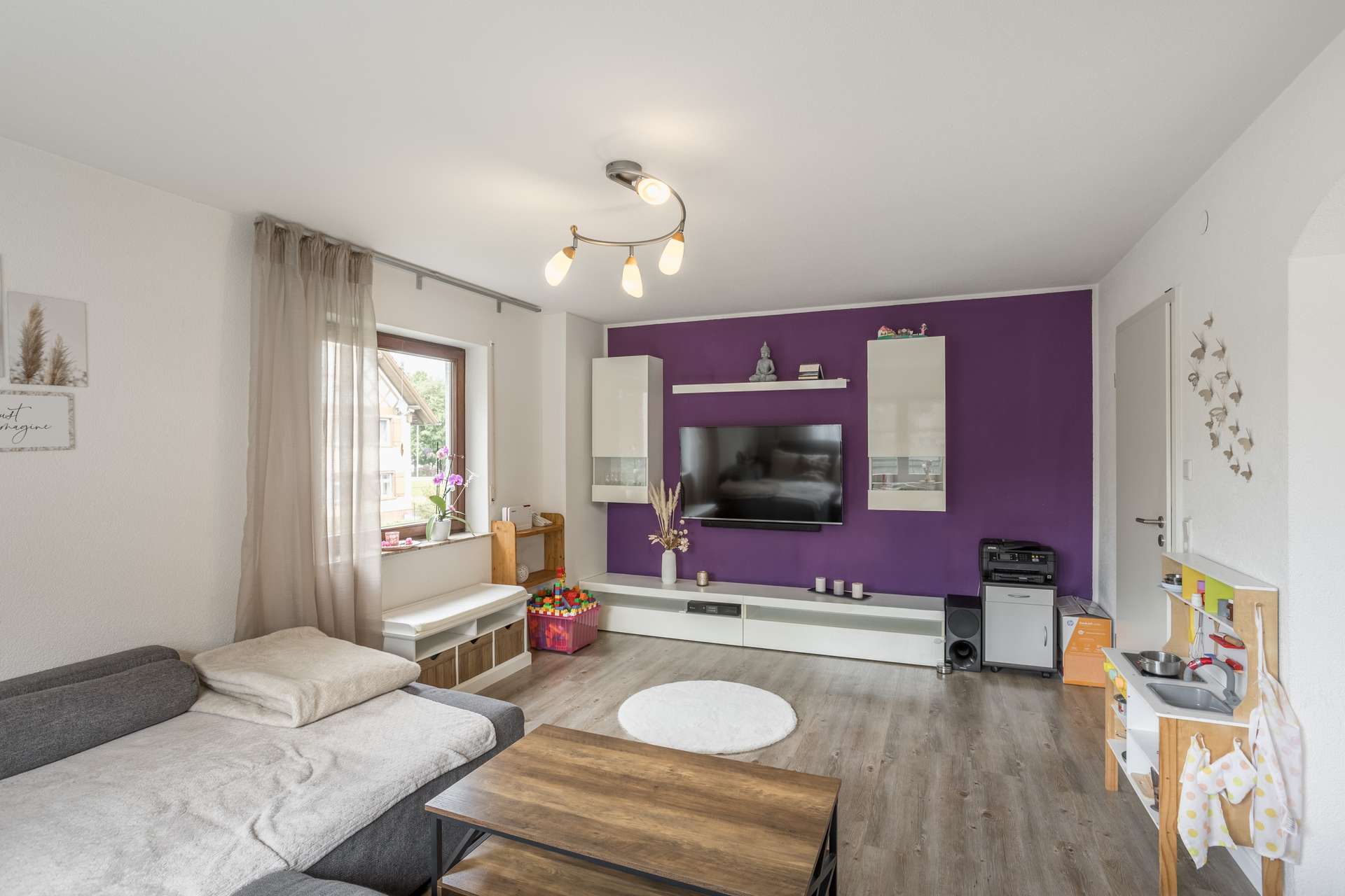 EG Ein Wohnzimmer mit einem grauen Sofa, einem an der Wand montierten Fernseher vor einer violetten Akzentwand, weißen Aufbewahrungsschränken und einer Spielküche aus Holz für Kinder.