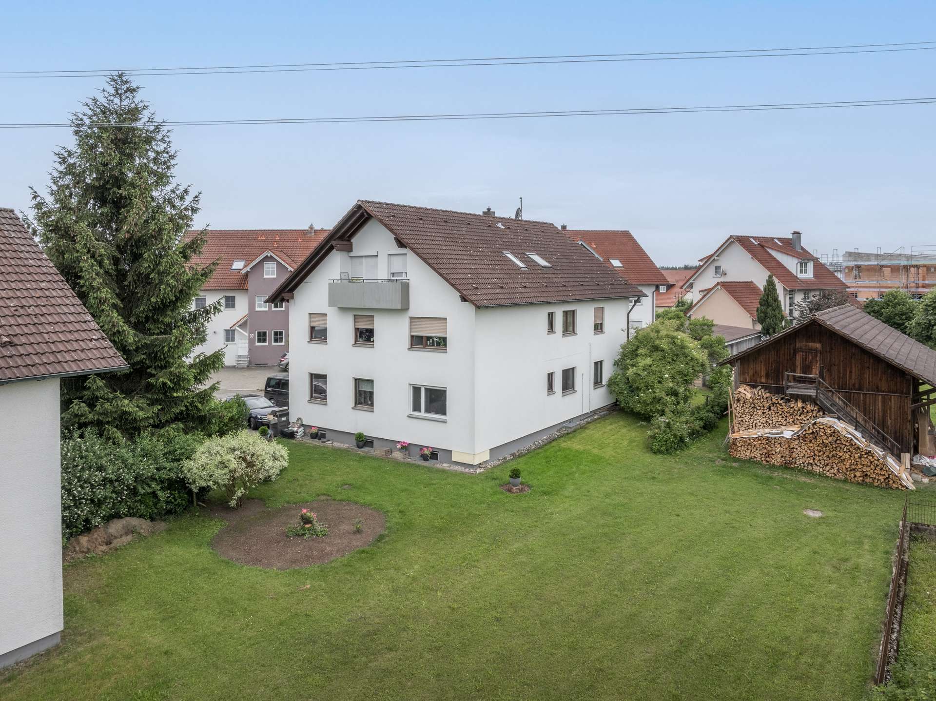 Außenansicht Hinter einem begrünten Vorgarten steht ein weißes, mehrstöckiges Wohnhaus mit einem braunen Ziegeldach; rechts davon befinden sich ein Holzschuppen und ein großer Holzstapel.